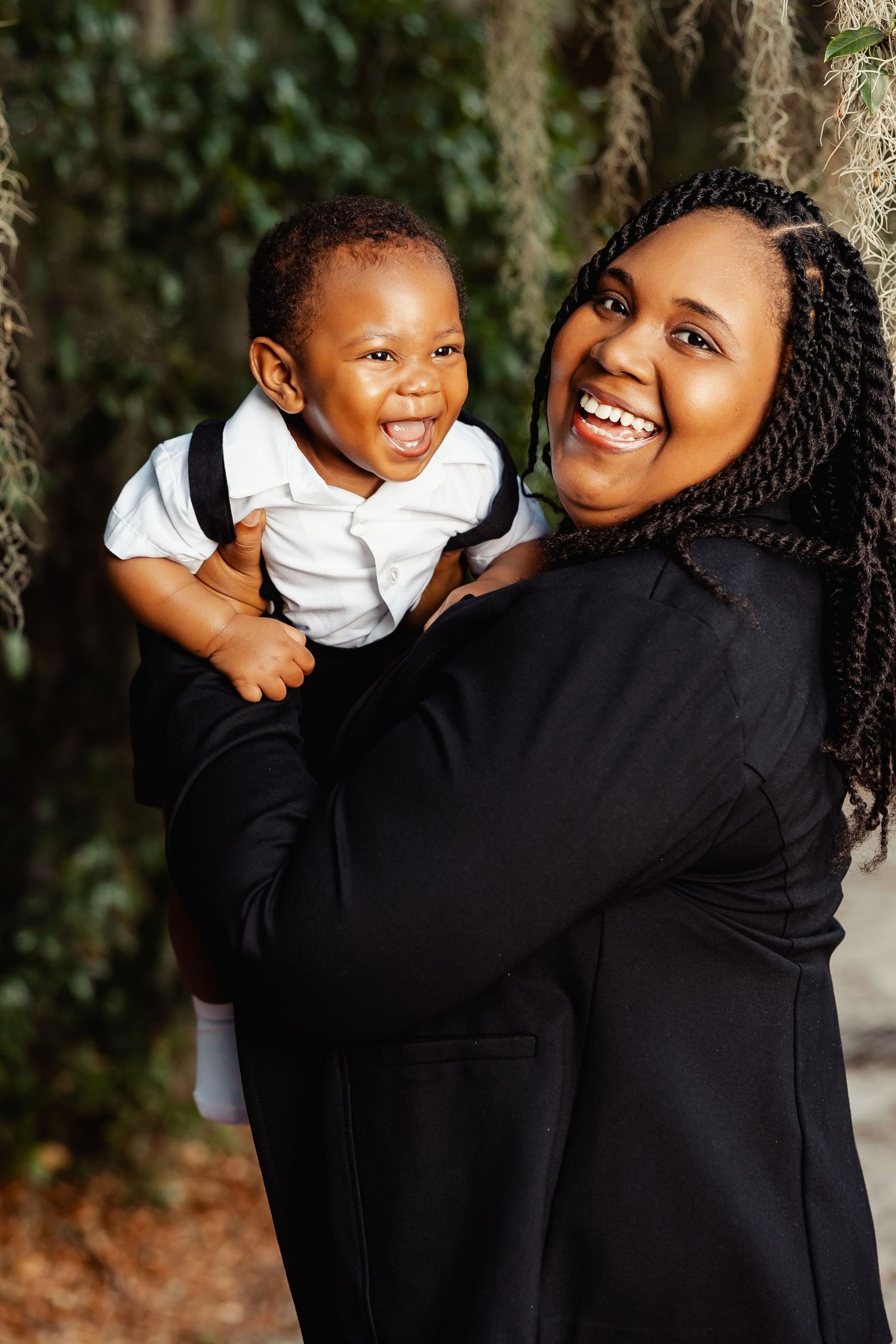 Happy Black mother holding laughing baby boy in white shirt and suspenders outdoors.