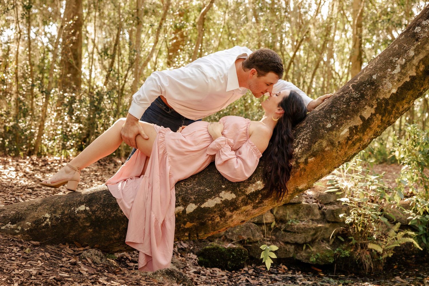 Romantic couple sharing a kiss outdoors, woman in flowing pink maternity dress draped over a mossy tree trunk.