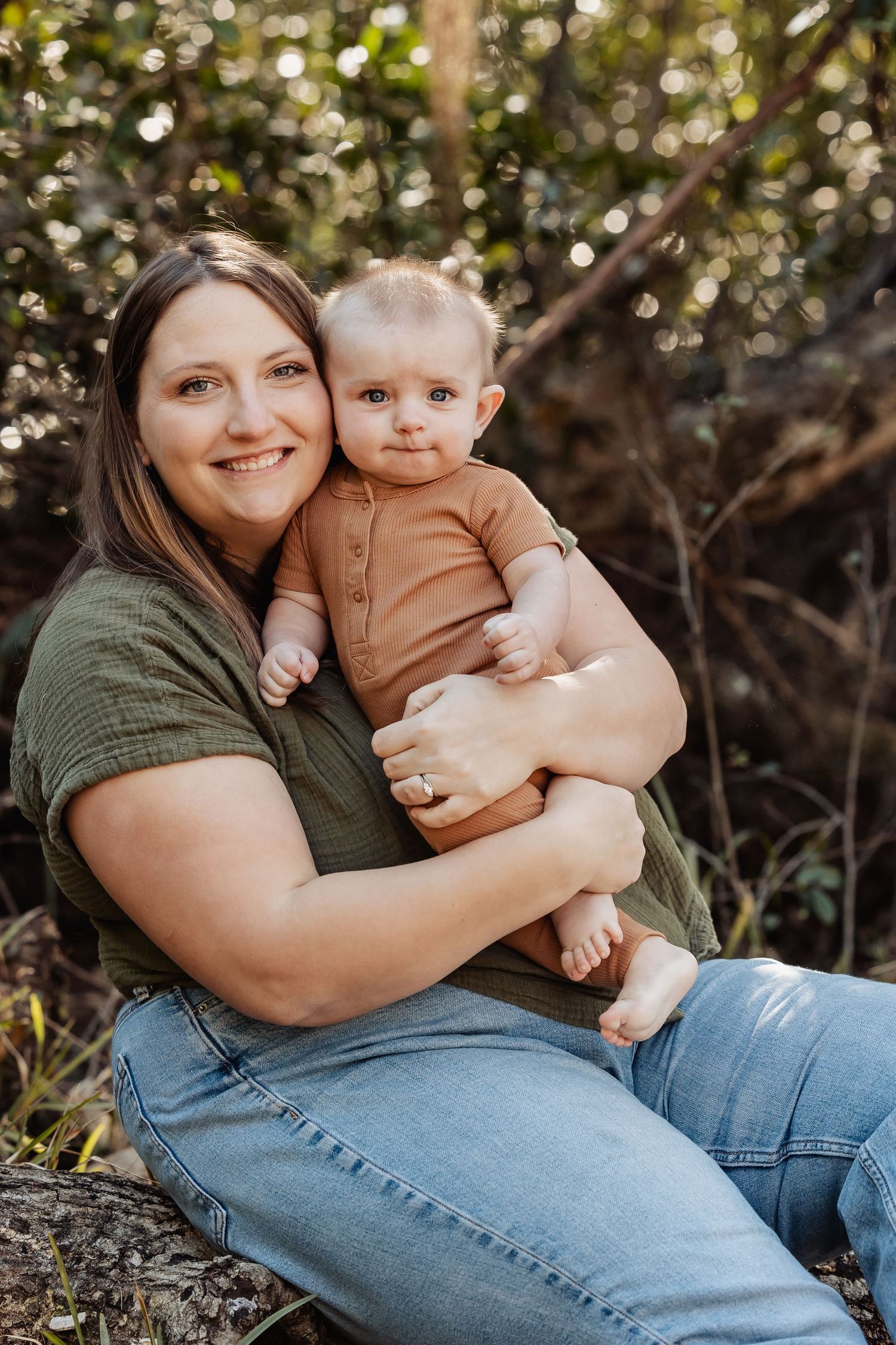 Smiling mother holding her baby boy while sitting outdoors in a natural green setting.