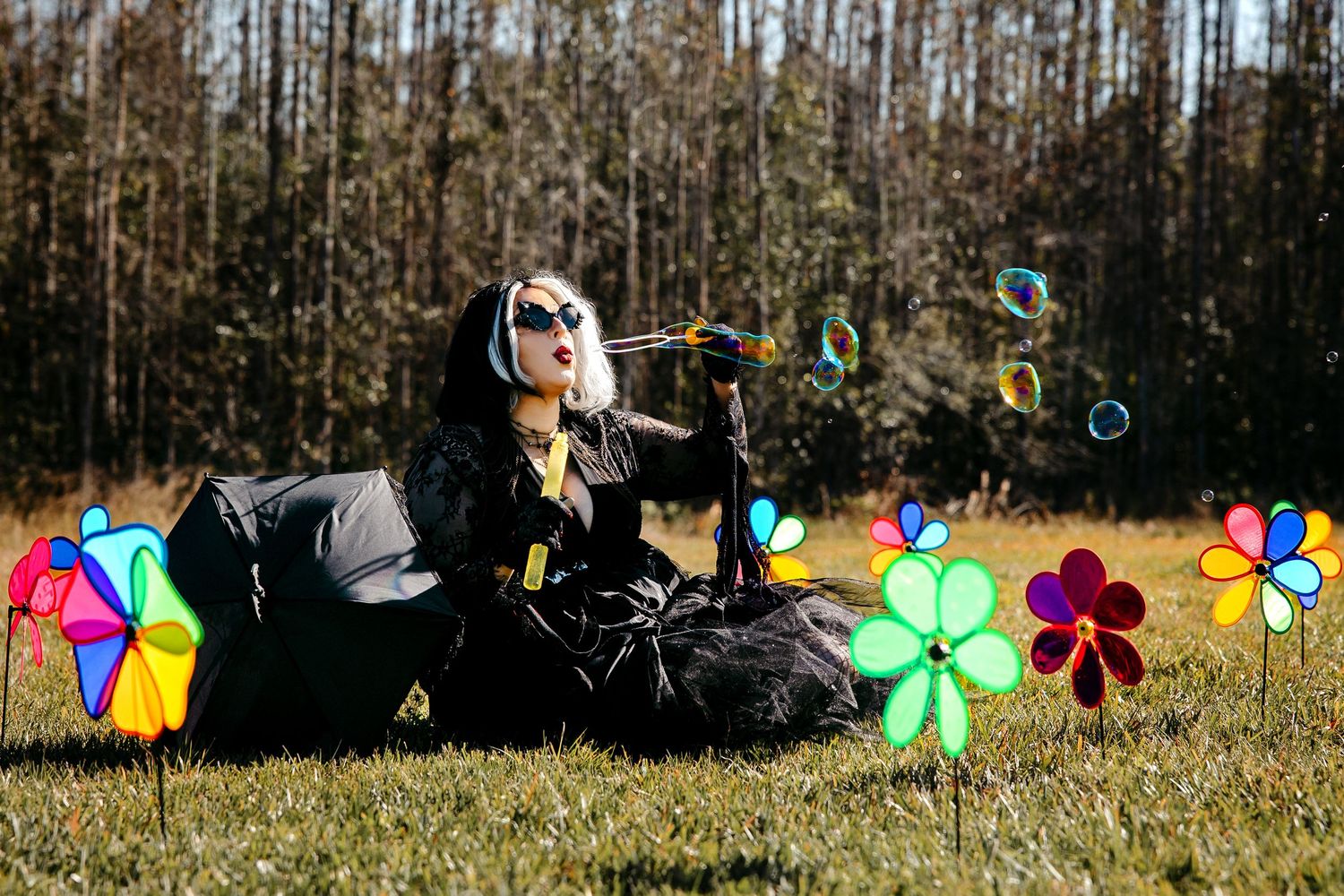 Woman in black outfit with white hair blows bubbles while lying on grass with colorful pinwheels.