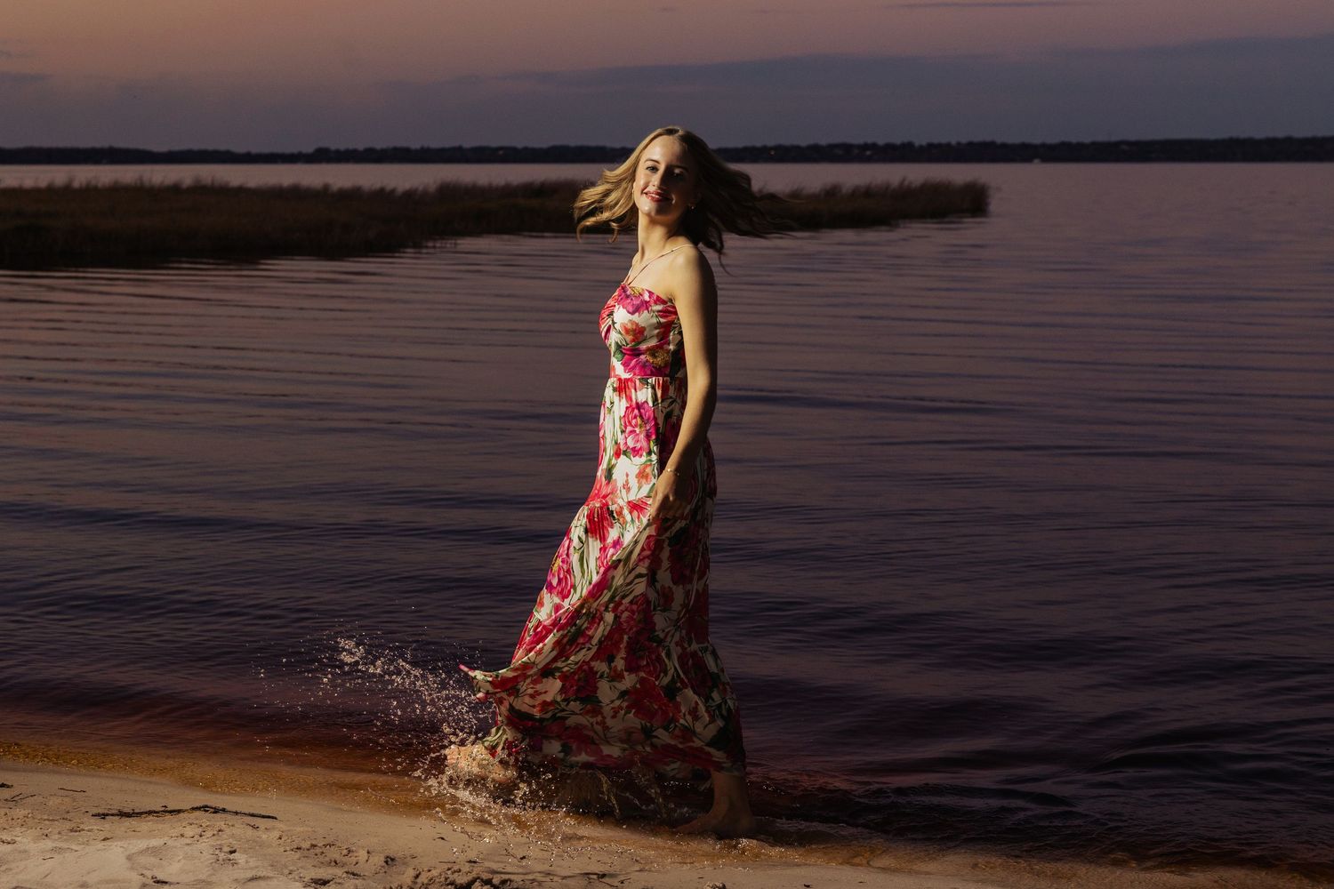 Woman in floral maxi dress standing in shallow water at sunset, hair flowing in the breeze.