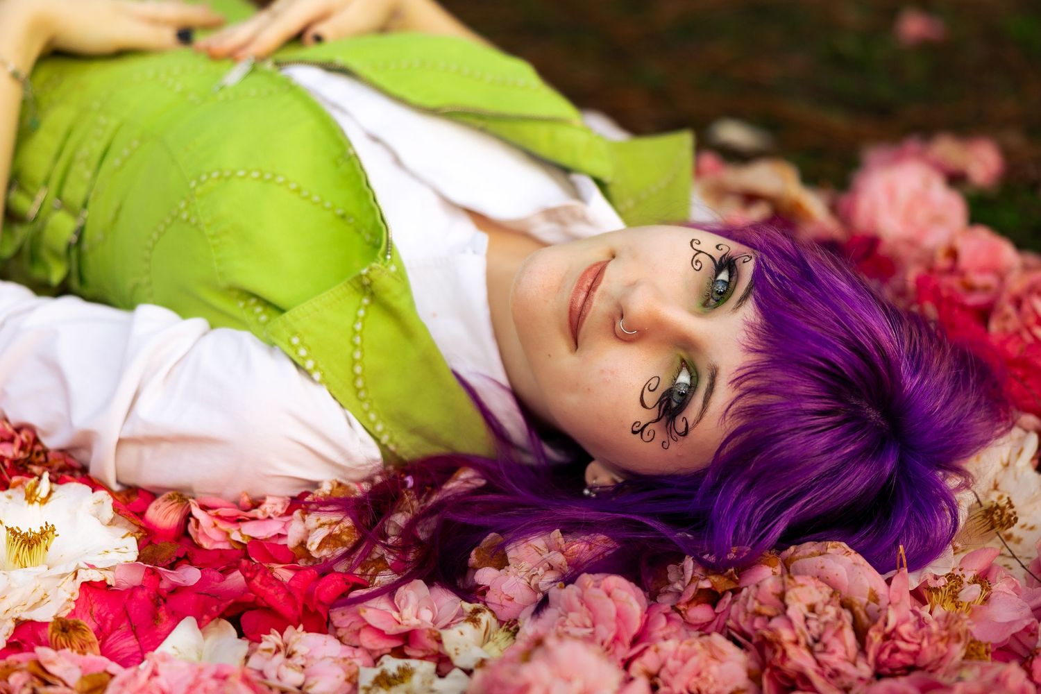 Woman with purple hair and green dress lying among pink flowers with butterfly eye makeup.