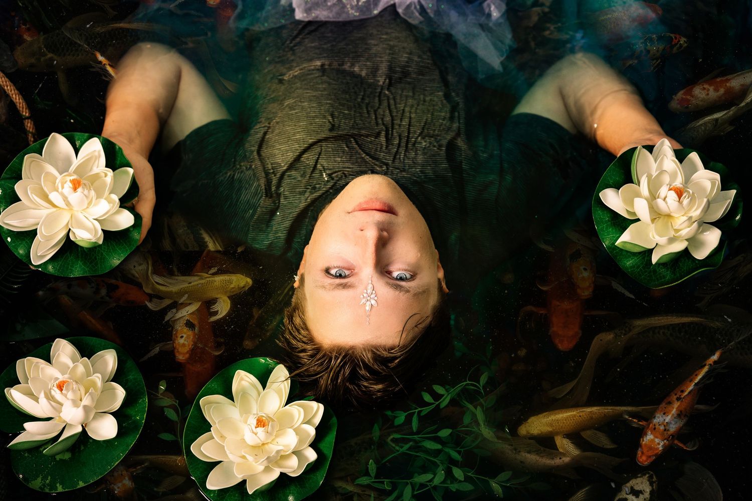 Overhead view of a person lying in water surrounded by white lotus flowers and koi fish.