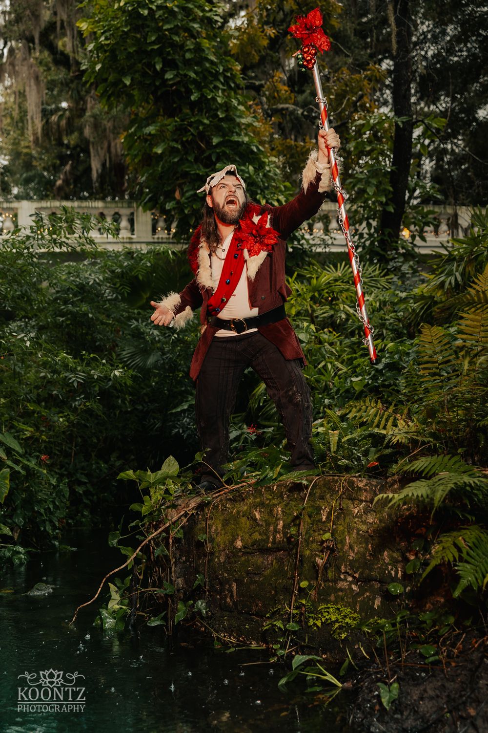 A cosplayer in a red coat dramatically raises a decorated red staff while standing on rocks near water in a lush garden.