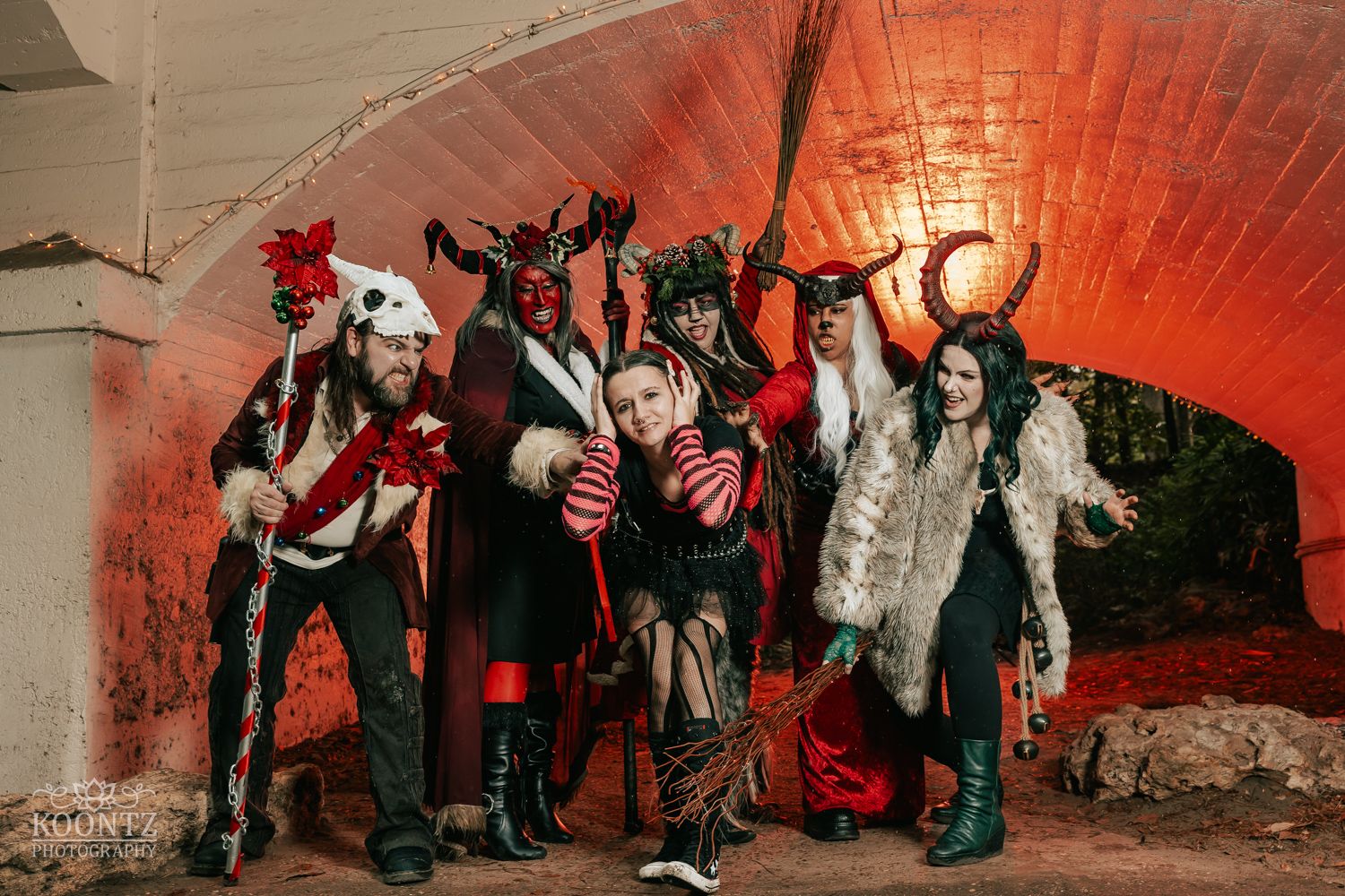 Six people in elaborate red and black devil costumes pose together under an arch with red glow.