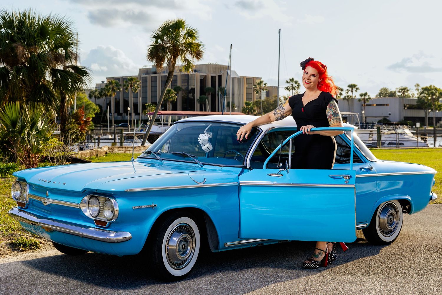 Tattooed redhead woman in black dress poses with classic blue vintage Corvair car outdoors.