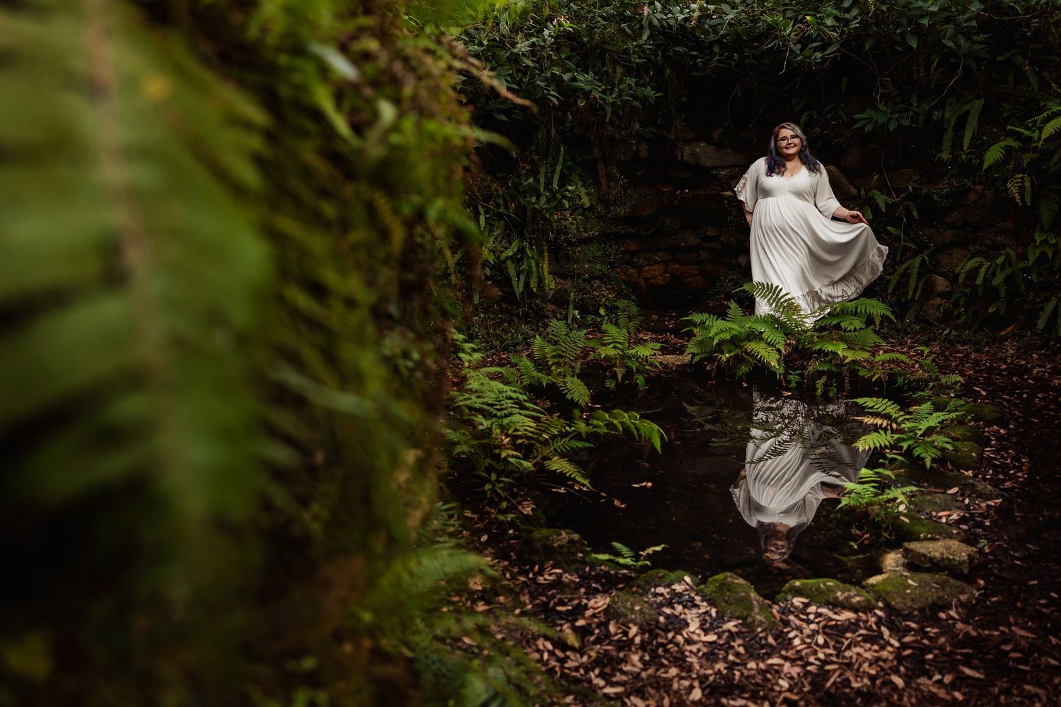 Bride in white gown standing by a reflective forest pond surrounded by lush ferns and greenery.
