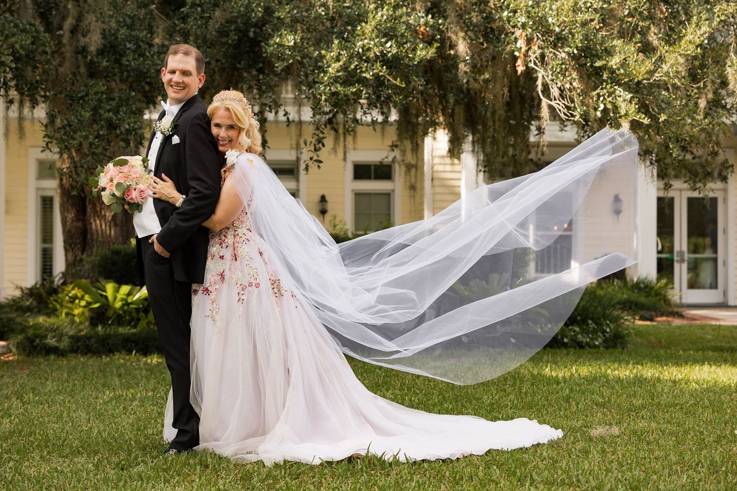 Bride and groom pose outdoors with flowing veil billowing in the wind near a historic home.
