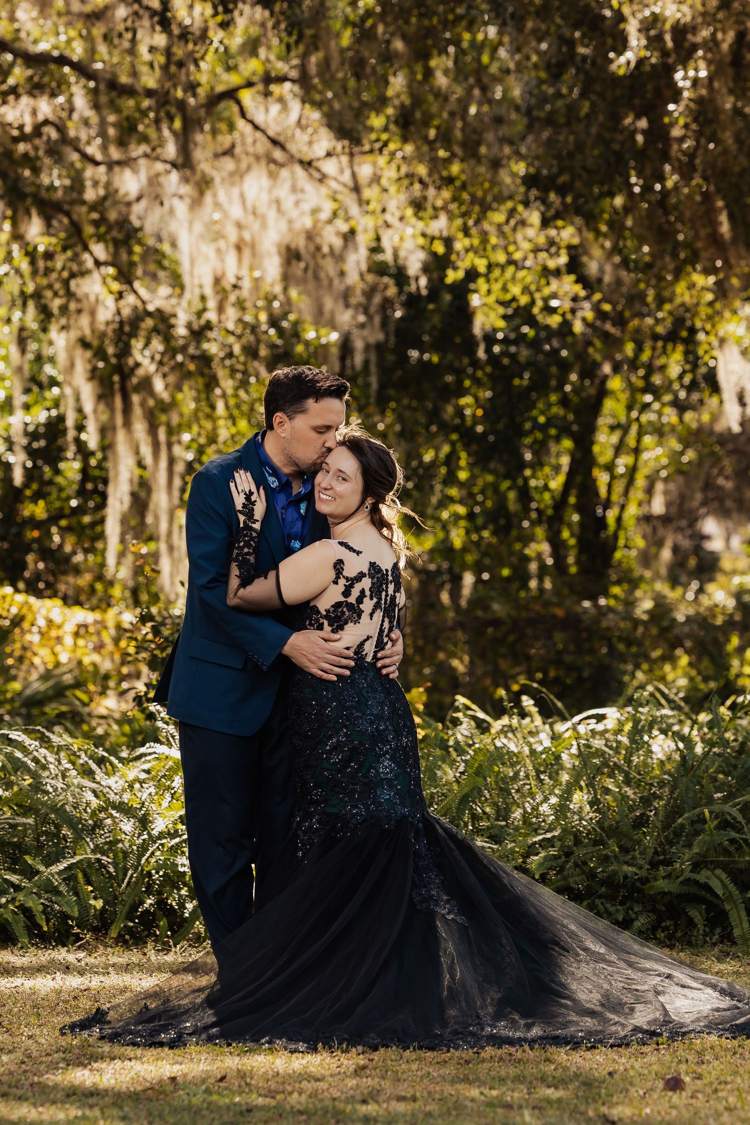 Couple embraces romantically under Spanish moss-draped trees, bride in stunning black mermaid gown at garden wedding.