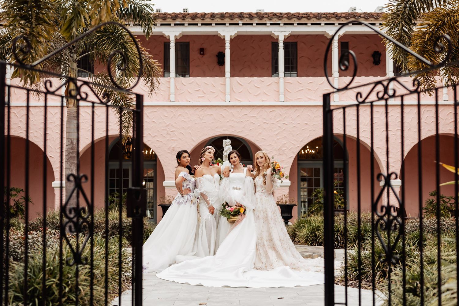 Three brides in white gowns posing through ornate iron gates at pink Mediterranean wedding venue.