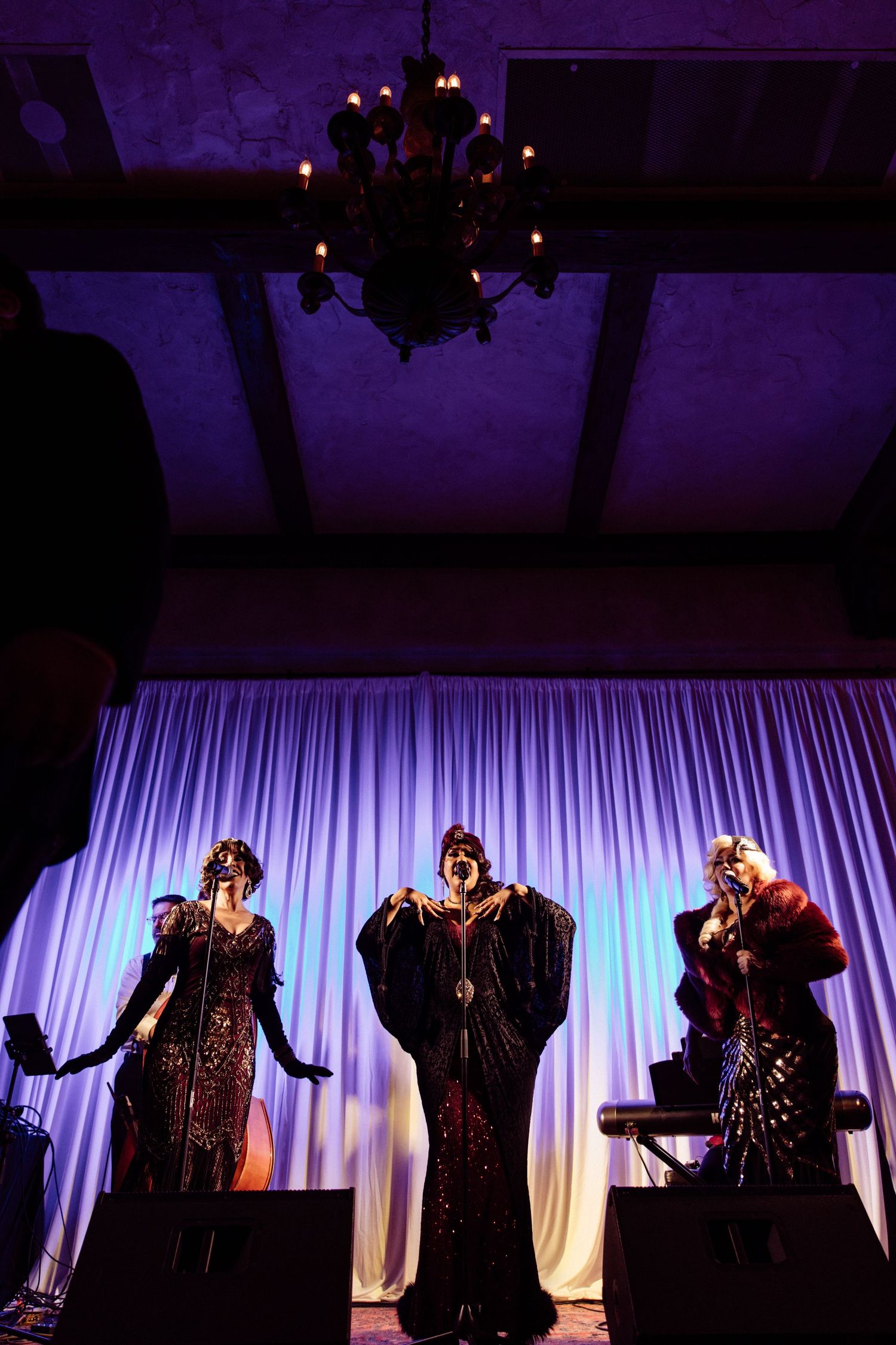 Three performers in glamorous vintage gowns singing on stage at wedding reception under chandelier.