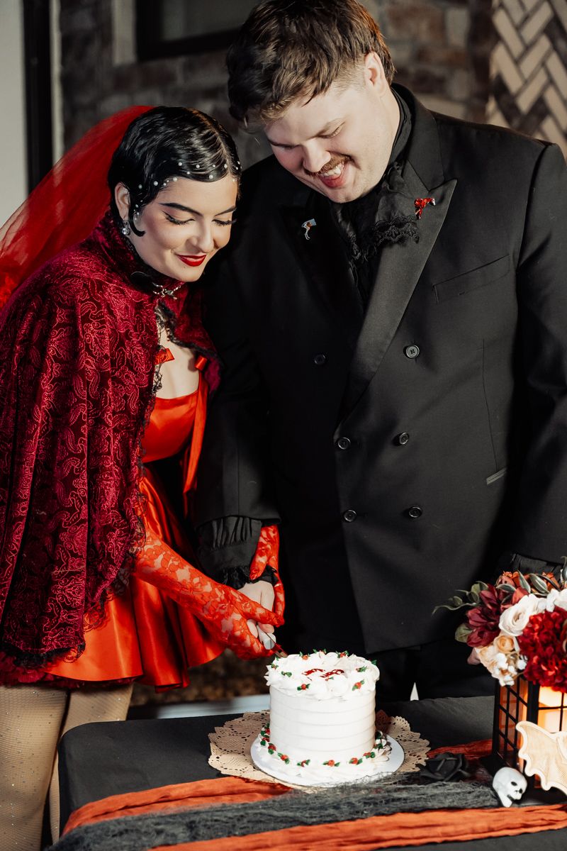 Bride in red lace cape and groom in black suit smile while cutting white wedding cake together.