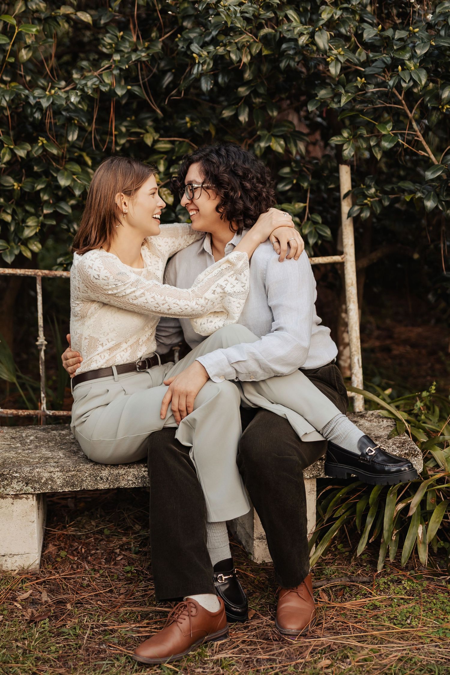 Two women sitting on a stone bench in a garden, laughing and embracing each other affectionately.