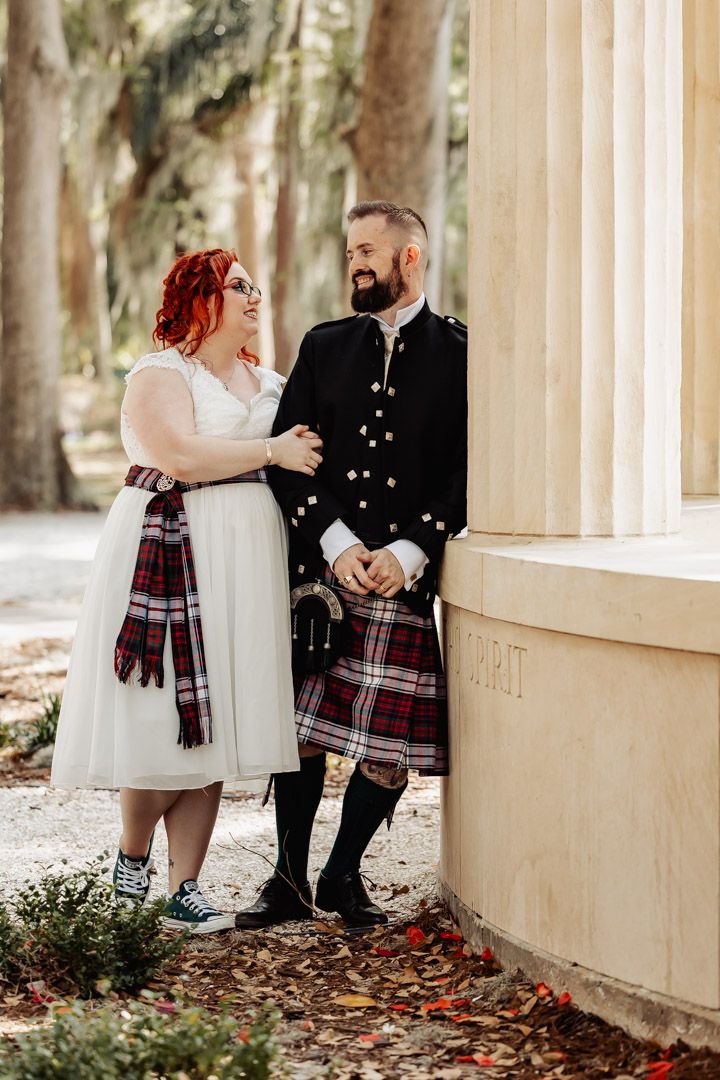 A couple in Scottish wedding attire stands together outdoors, with the bride wearing a white tea-length dress and blue sneakers.