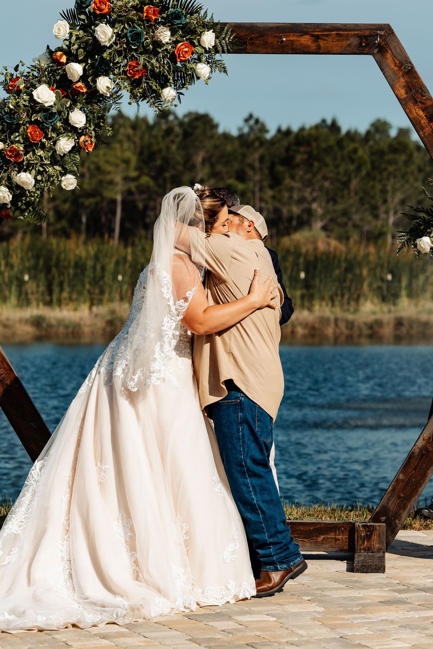 Romantic moment under floral-decorated wooden arch by lakeside during sunset ceremony.