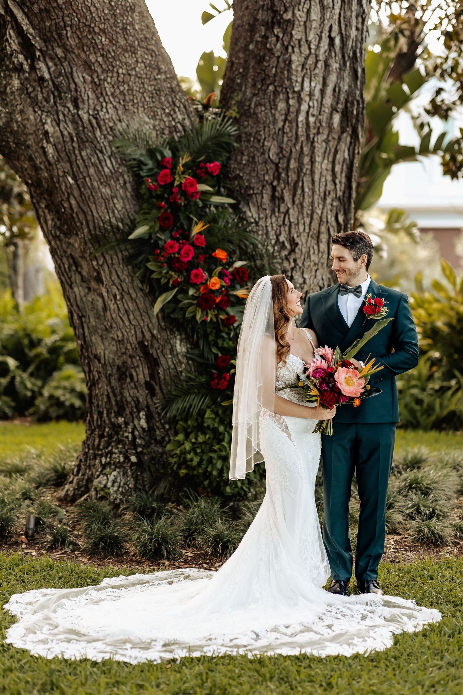 Bride and groom pose together by large tree with vibrant tropical red and pink floral arrangement.