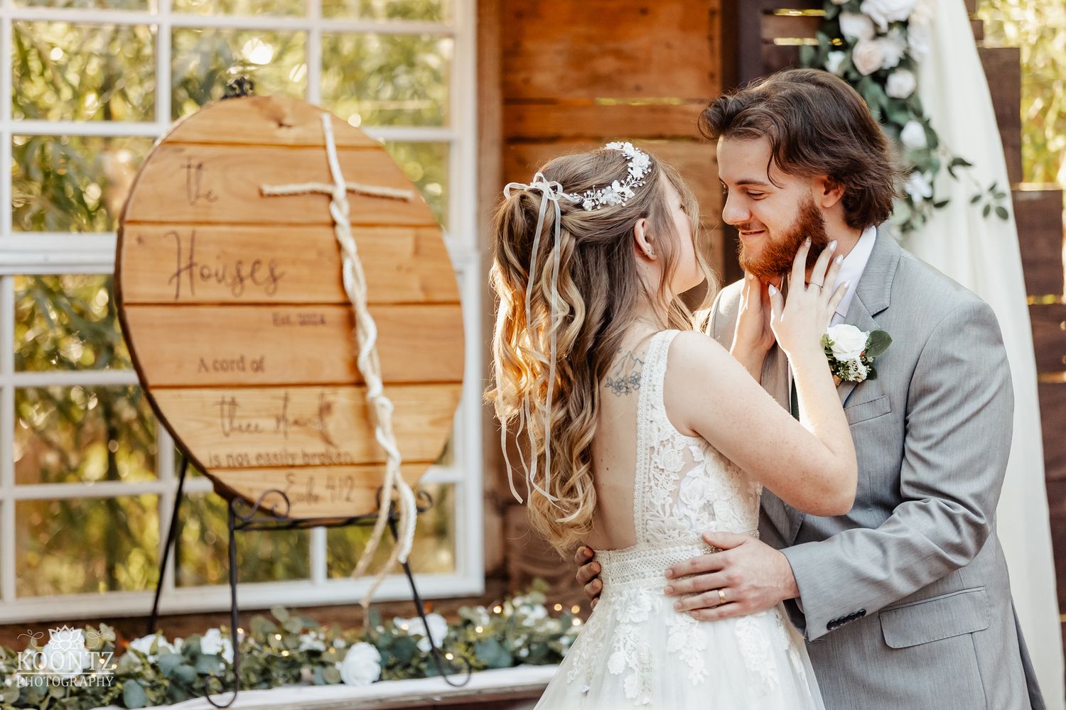 Bride and groom share romantic moment near wooden circular wedding sign at rustic outdoor venue.