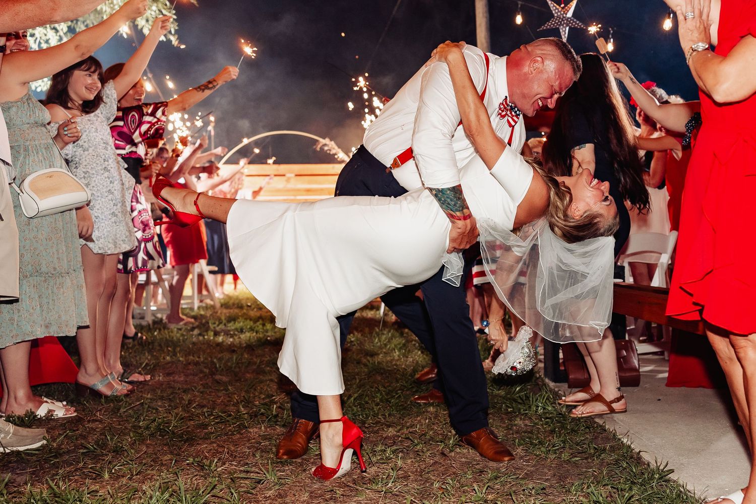Groom dips bride dramatically during sparkler send-off exit at outdoor evening wedding celebration with guests watching.