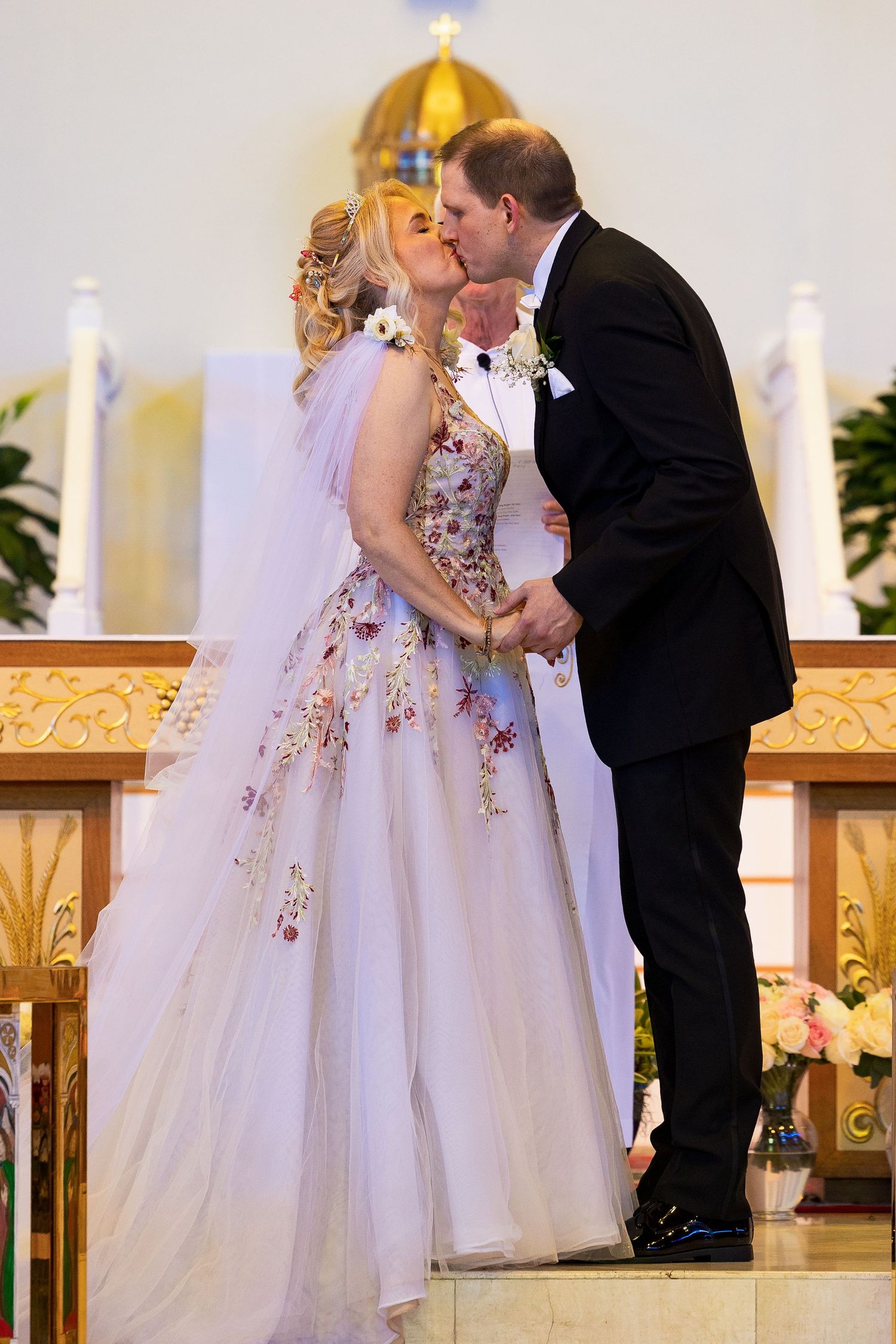 Bride and groom share first kiss at church altar, bride wearing floral embroidered wedding gown.