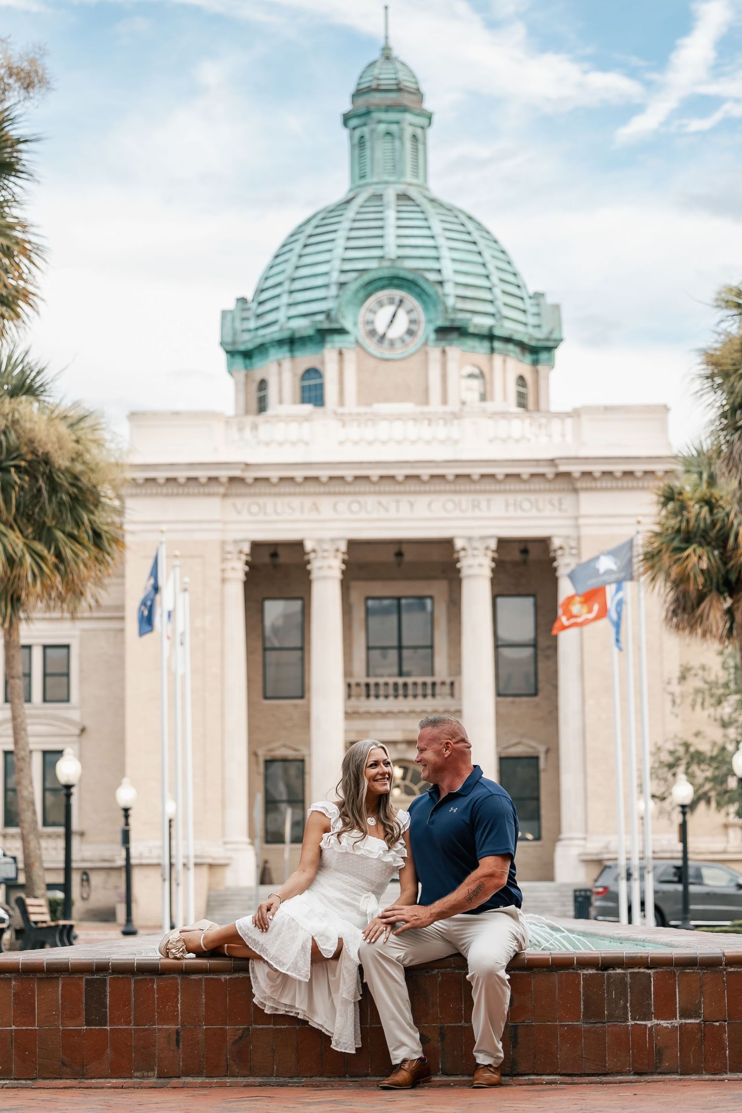 Engaged couple sitting together in front of Volusia County Courthouse with green dome and palm trees.
