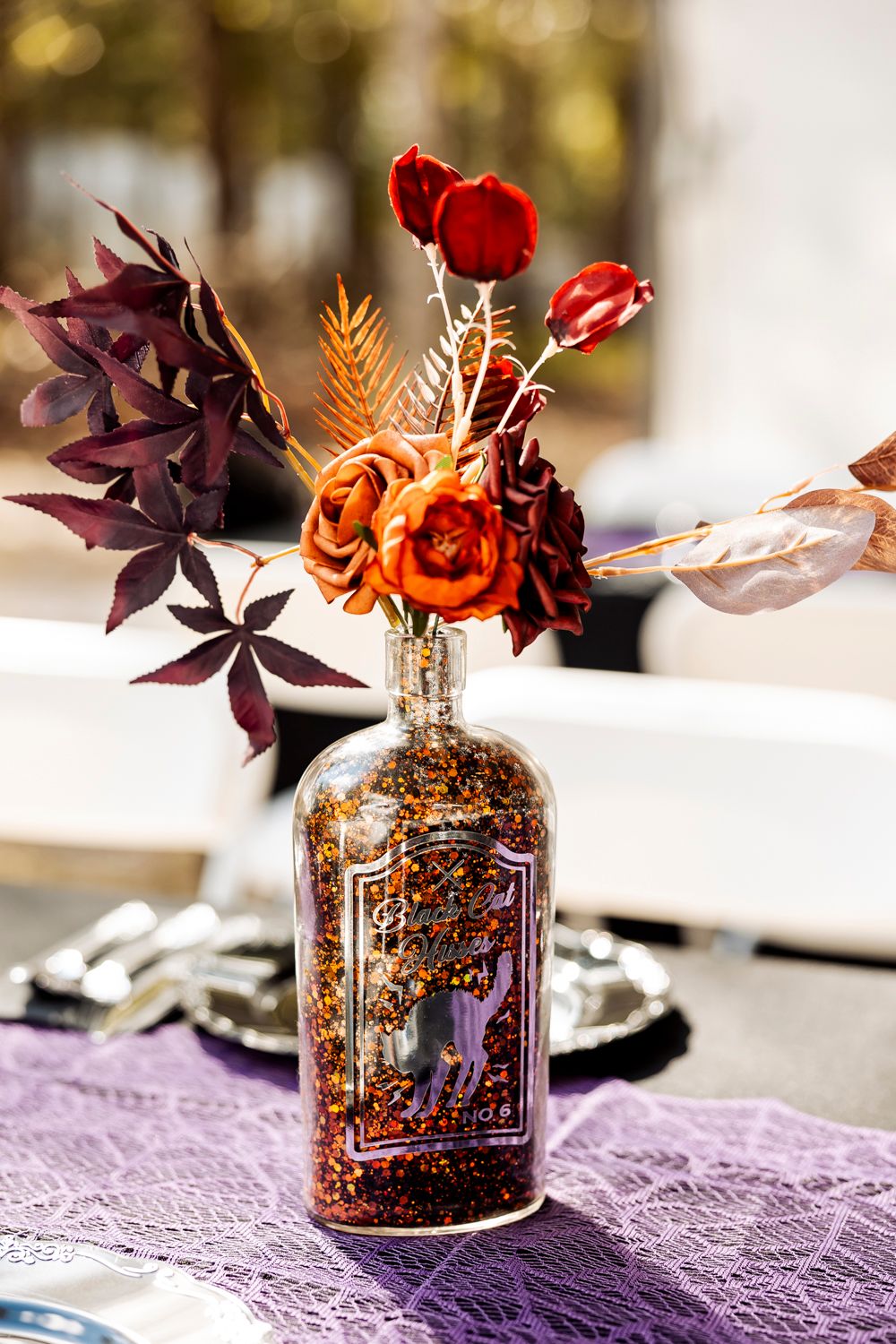 Decorative bottle filled with glitter holding red and burgundy autumn flowers on purple lace tablecloth.