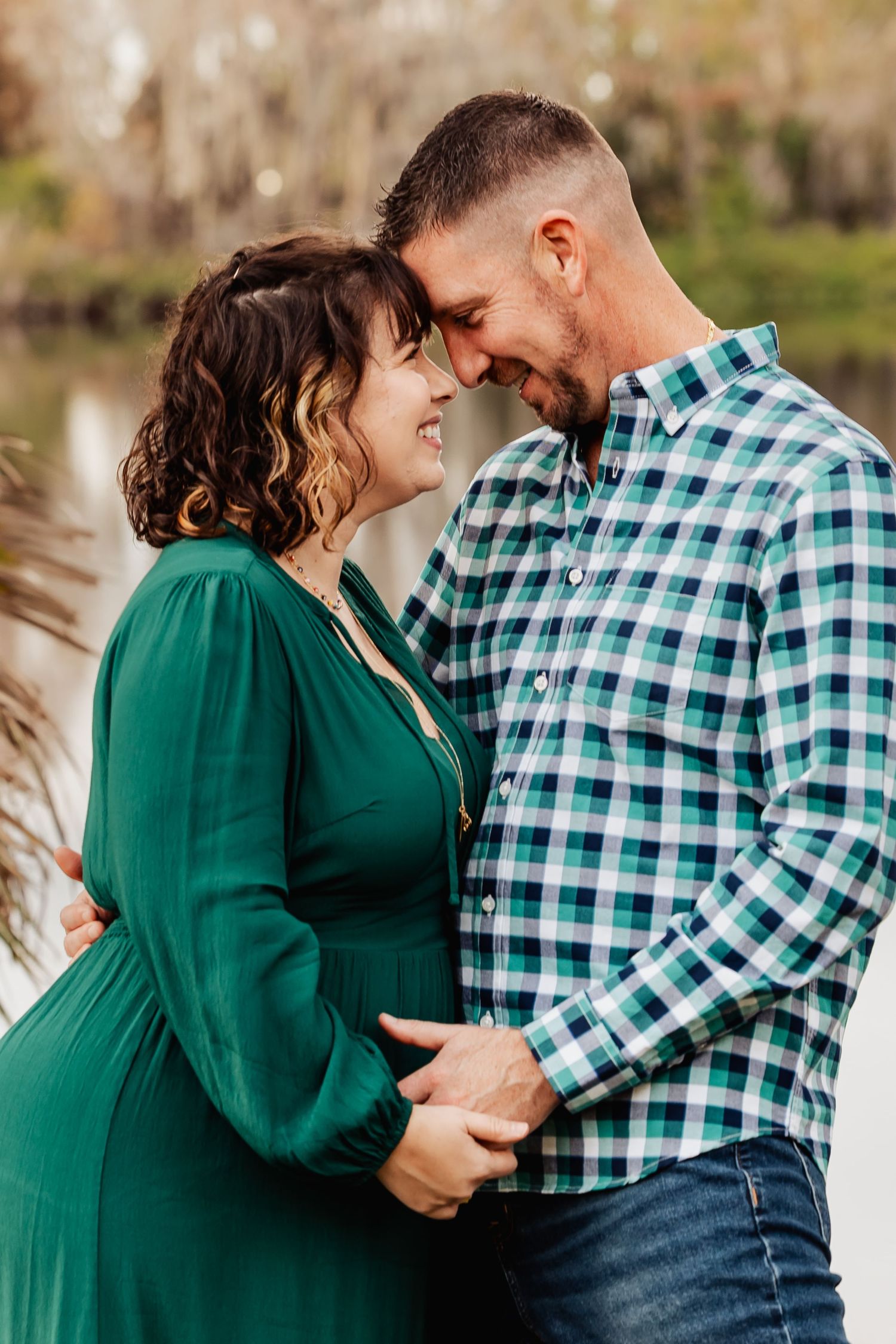 Pregnant couple in green dress and plaid shirt sharing tender forehead touch outdoors by water.