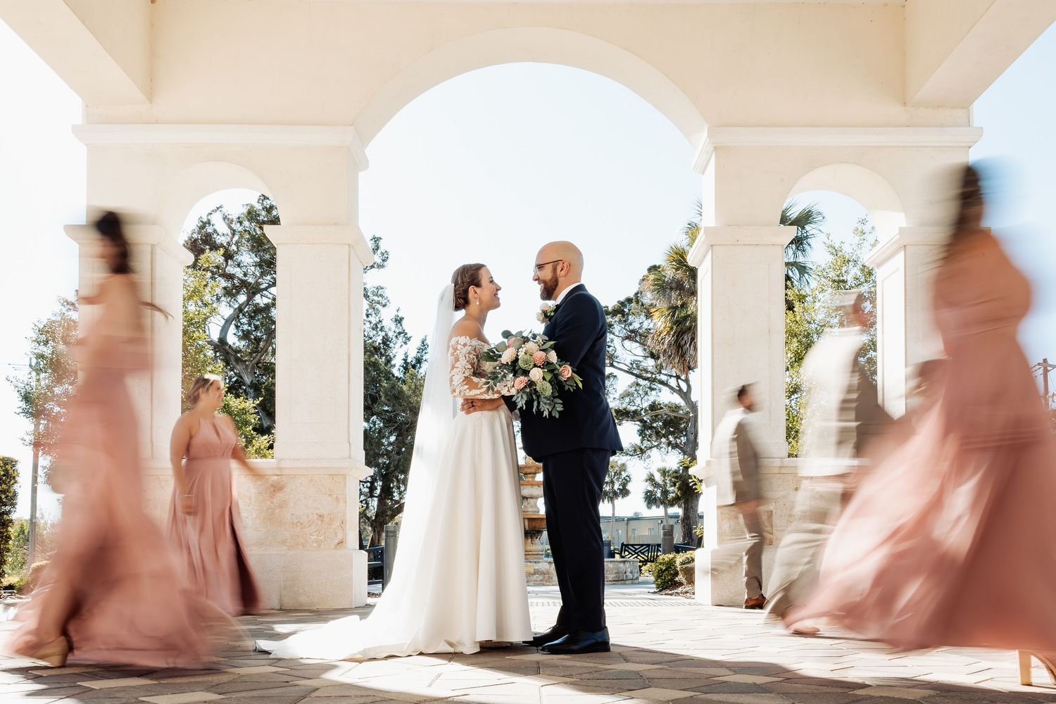 Bride and groom stand under white columns while wedding party in pink and tan attire moves past them.
