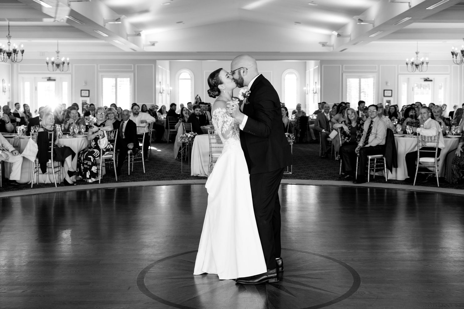 Bride and groom share first dance in black and white photo with wedding guests seated in background.