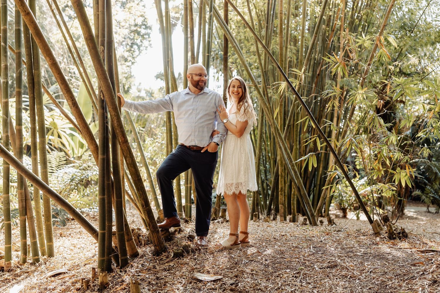 Couple posing among tall bamboo trees in a lush garden, woman in white dress and man in casual attire.