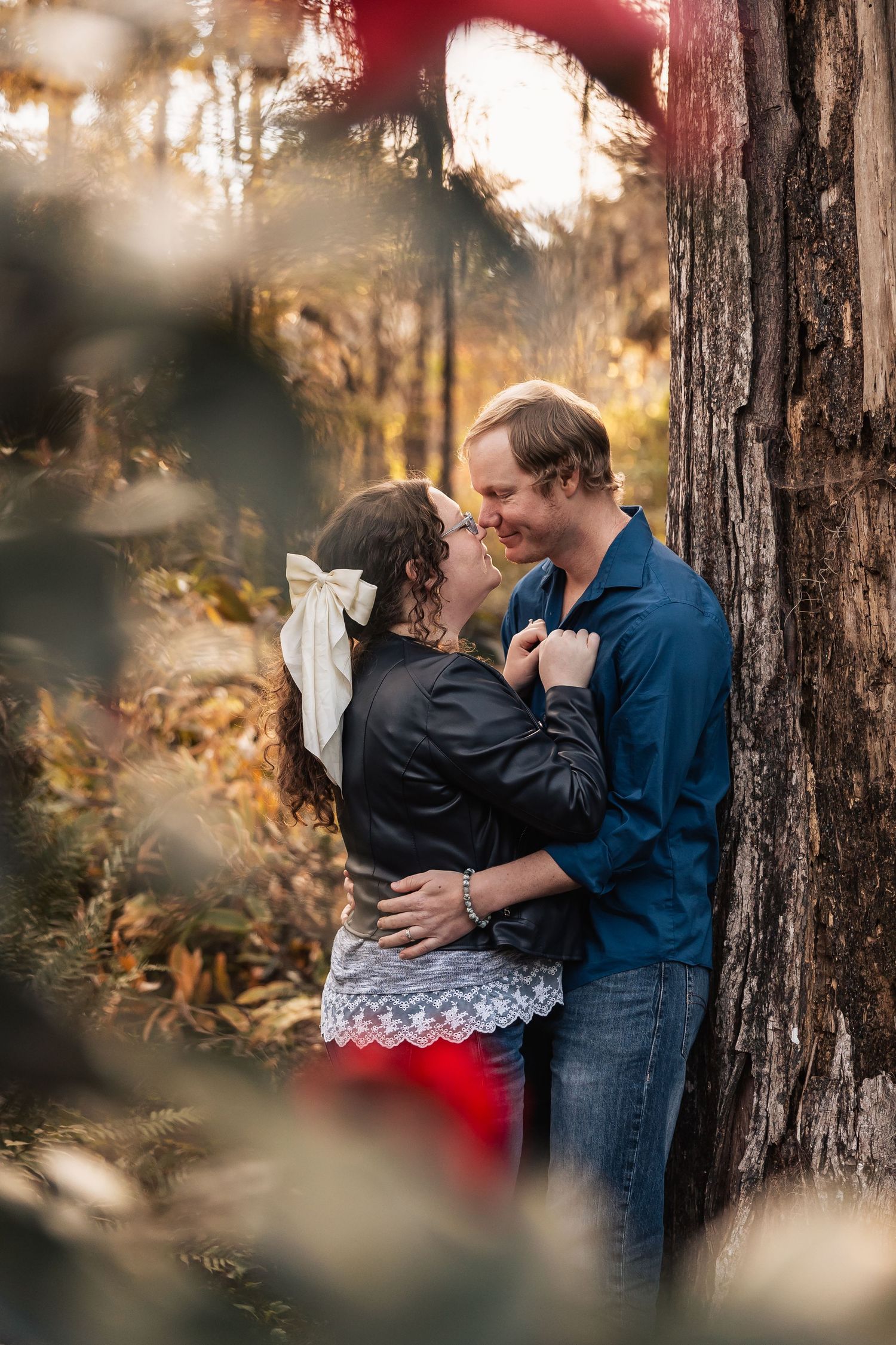 Couple embracing and smiling at each other leaning against a tree in autumn woods.