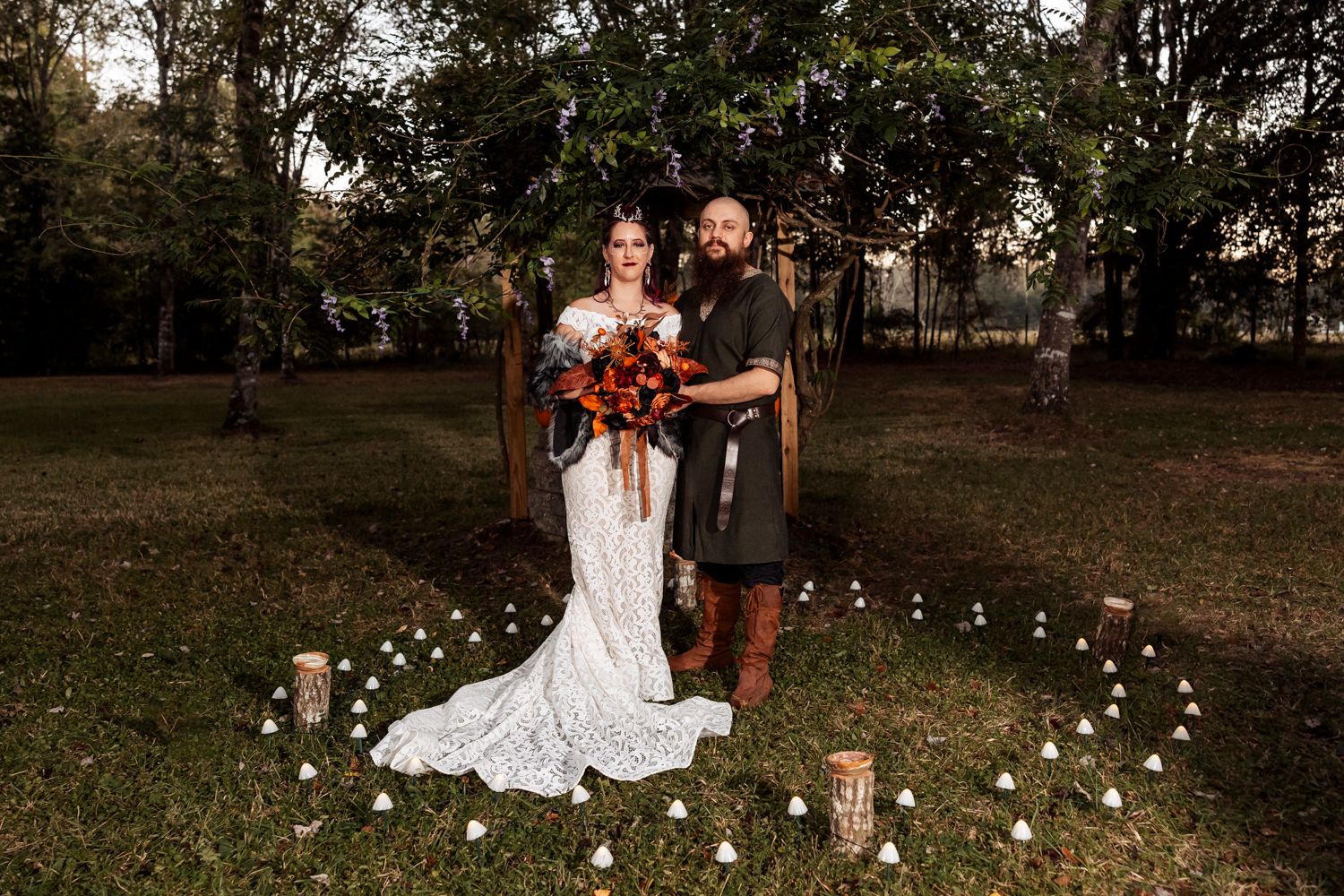 Viking wedding couple poses under rustic arch surrounded by white mushroom decorations and fall foliage.
