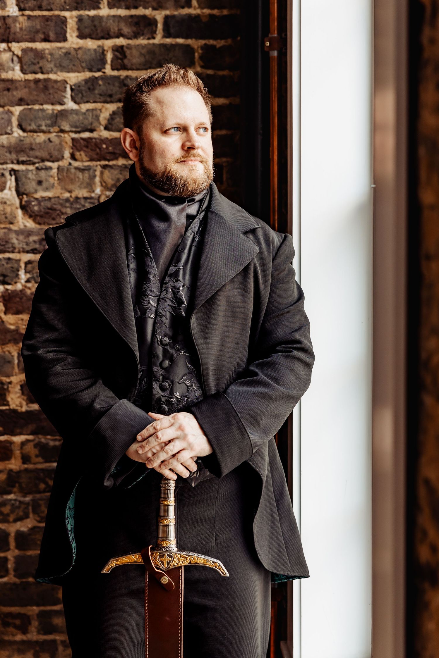 Bearded groom in black Victorian coat holding ornate sword standing by brick wall window in dramatic portrait.