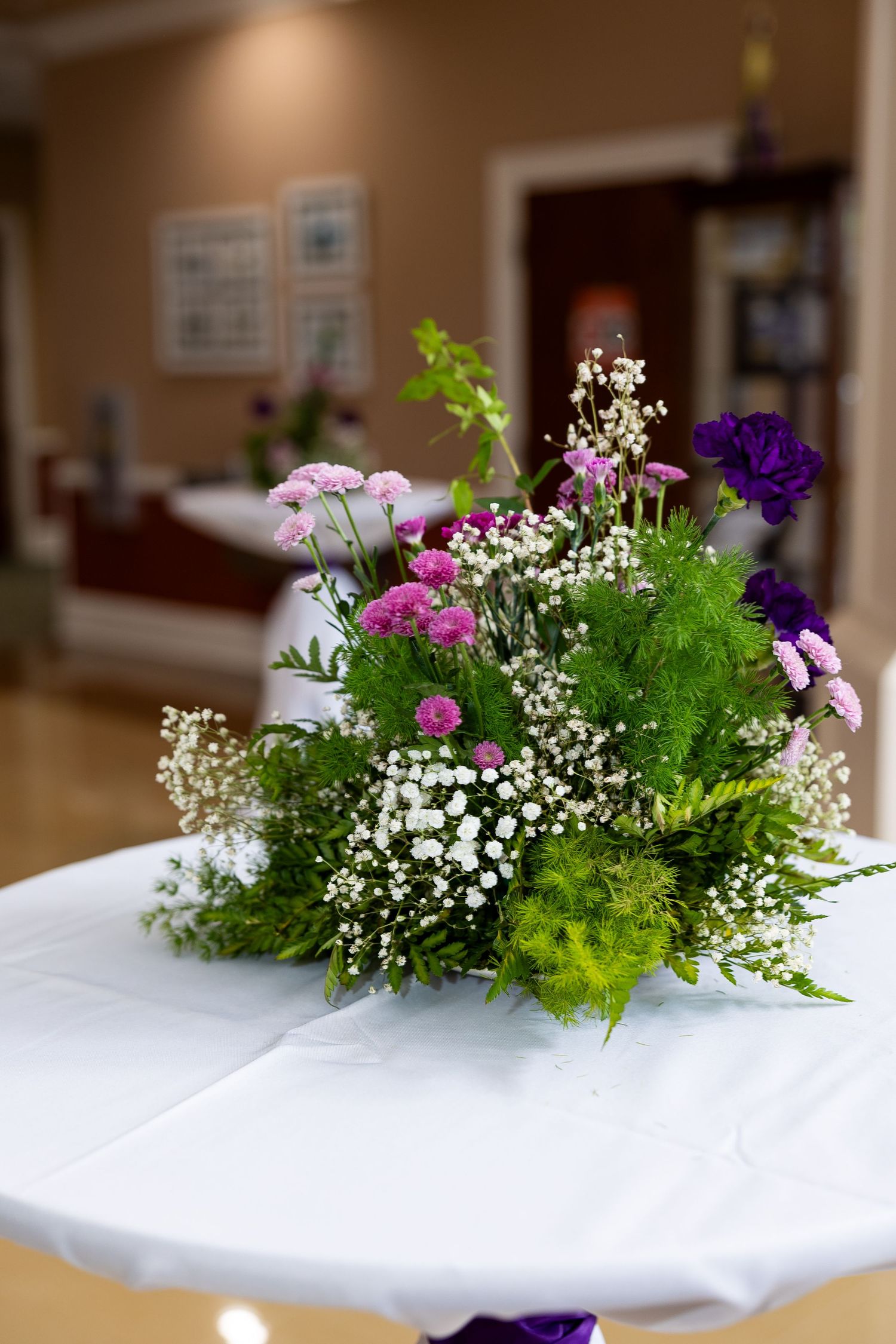 Beautiful floral centerpiece with pink, purple, and white flowers on a white tablecloth at an indoor event.