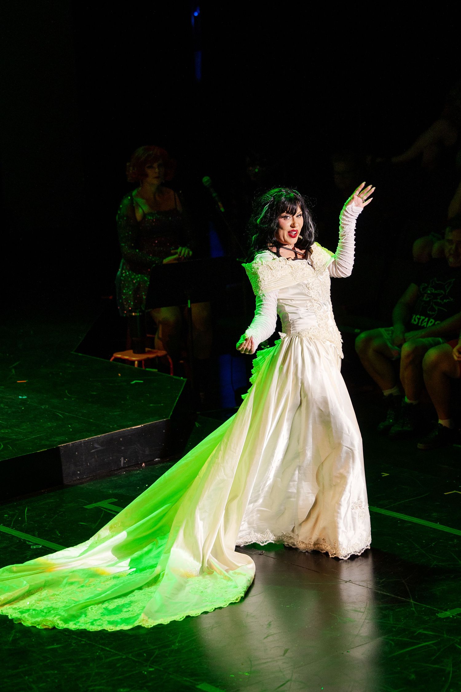 A performer in a white gown with a glowing green train gestures dramatically on a darkened cabaret stage.