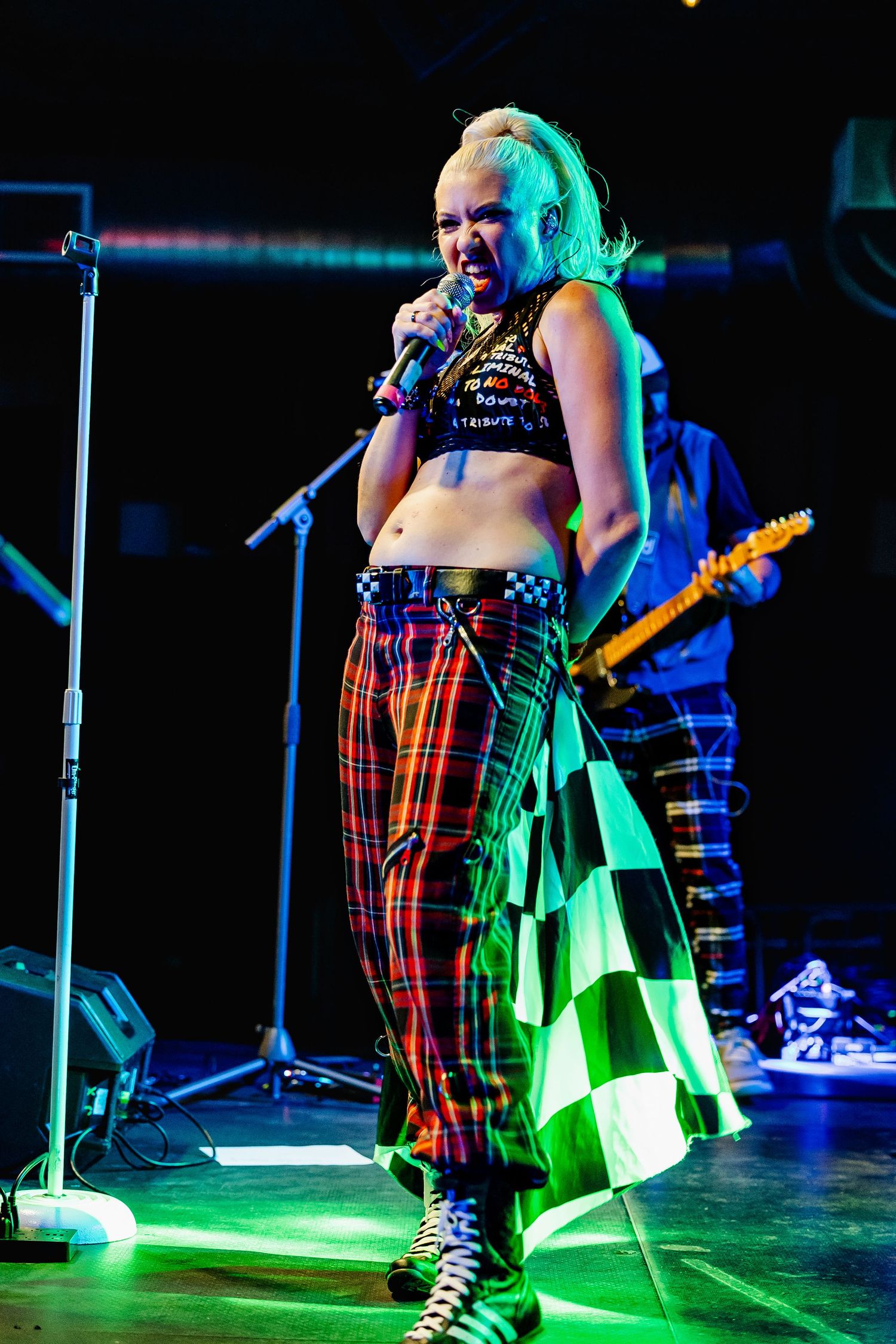 Female punk rock singer in plaid pants and crop top performs under green stage lights with microphone.