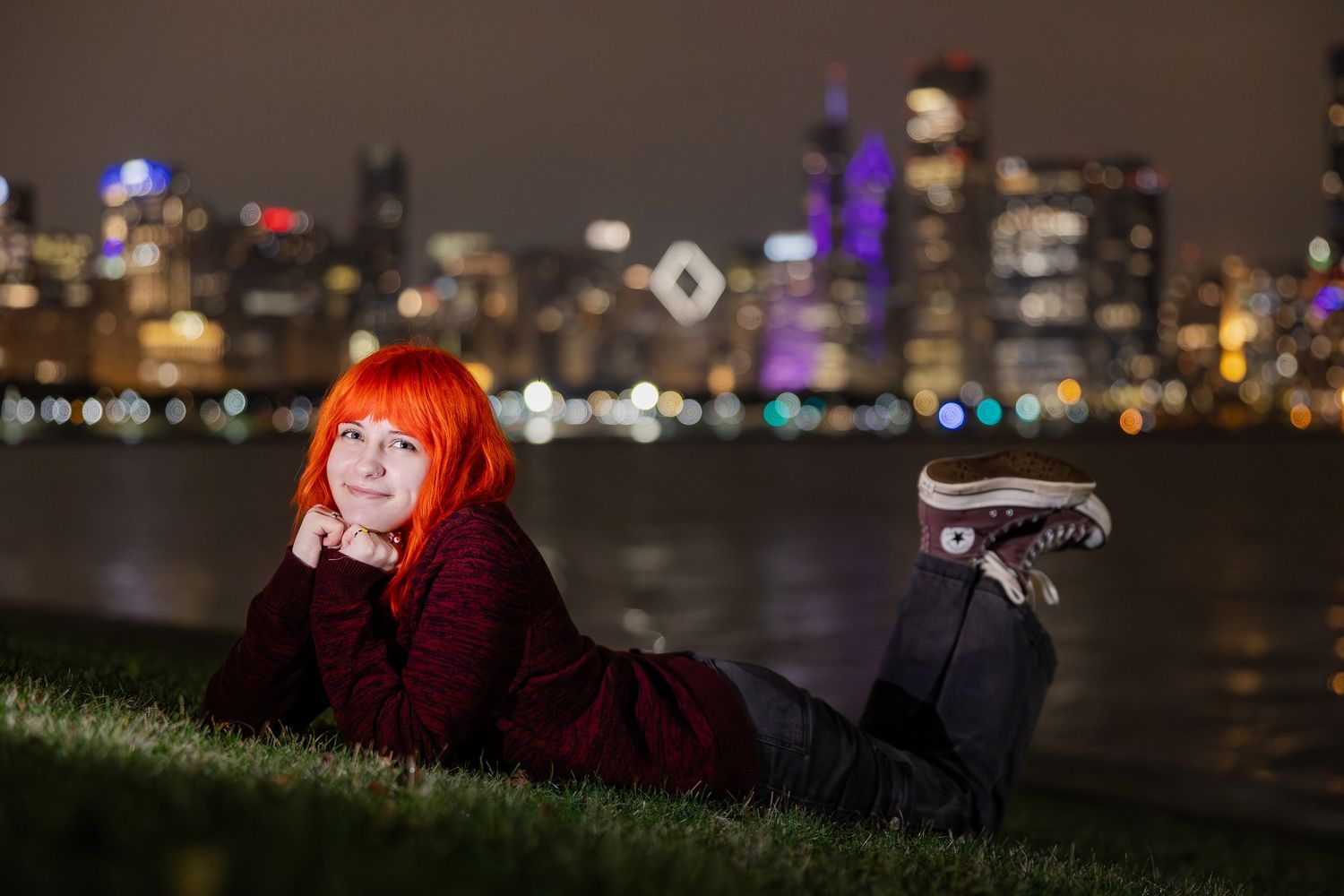 Red-haired woman lying on grass at night with illuminated city skyline and waterfront bokeh lights behind her.
