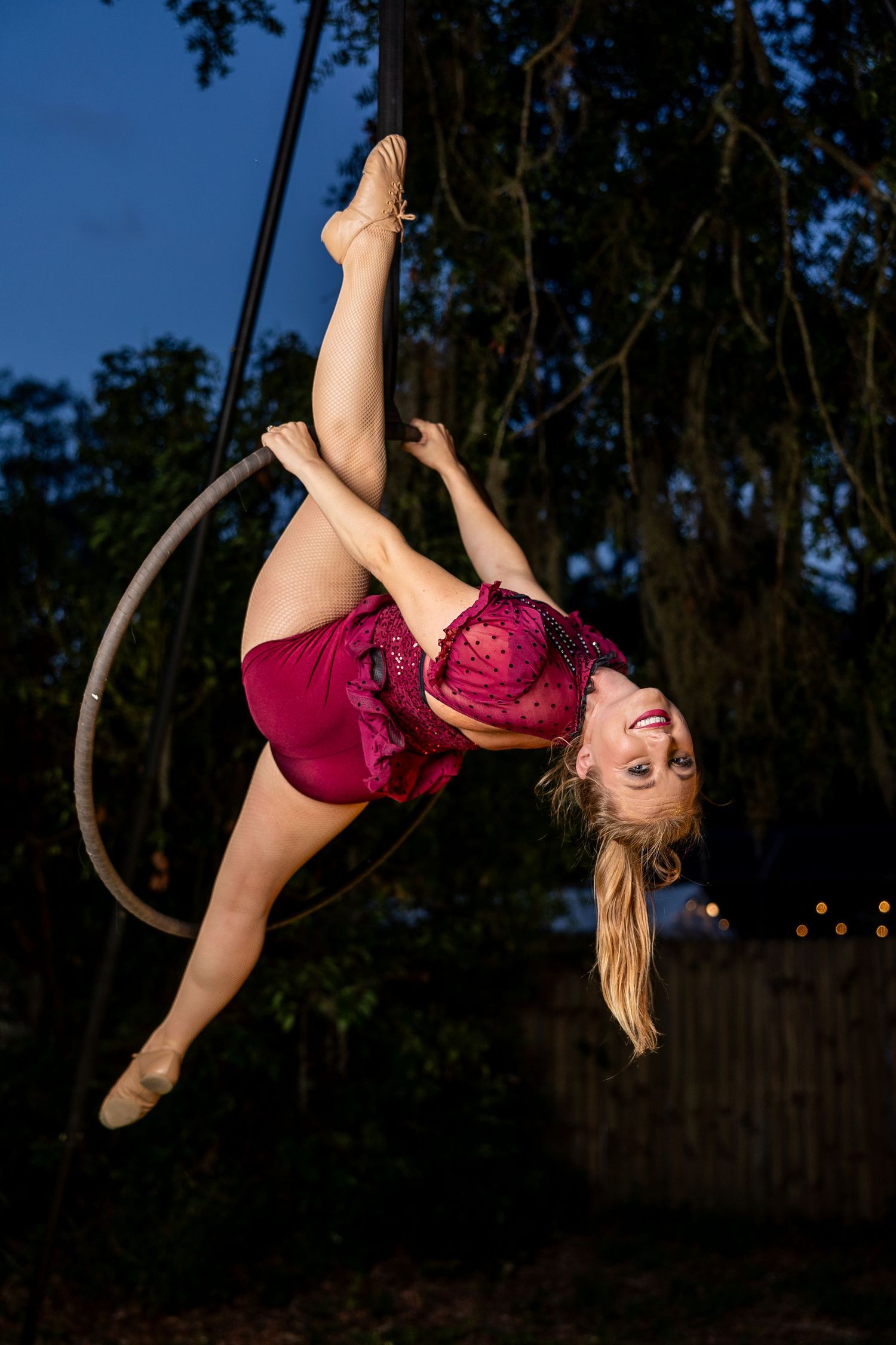 Smiling aerial hoop performer in red costume executing an inverted pose on a lyra at night outdoors.