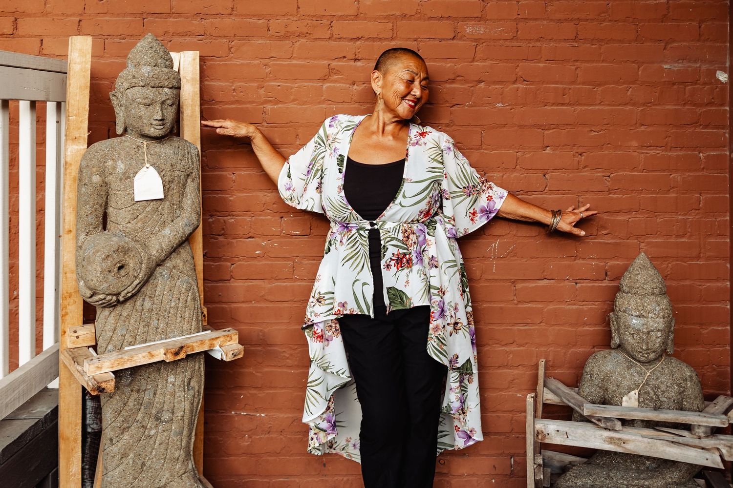 Confident woman in floral kimono poses between Buddha statues against a rustic brick wall outdoors.