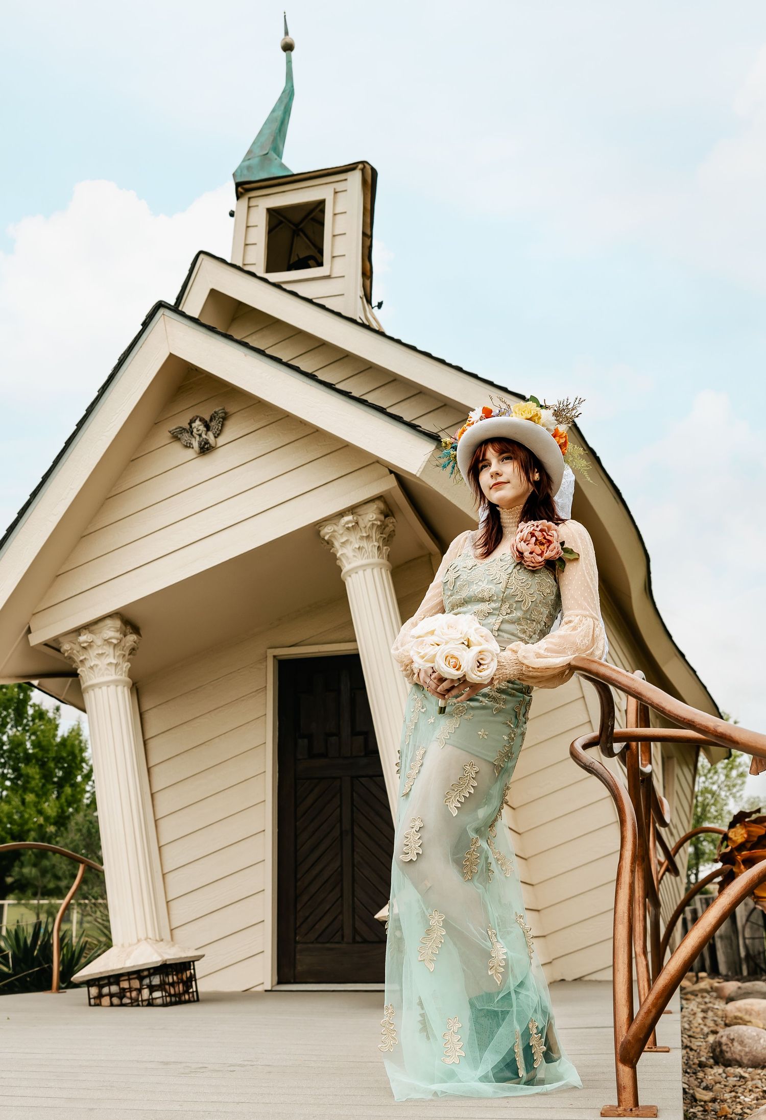 Bride in mint lace gown and floral hat poses with bouquet outside quaint white chapel under cloudy sky.