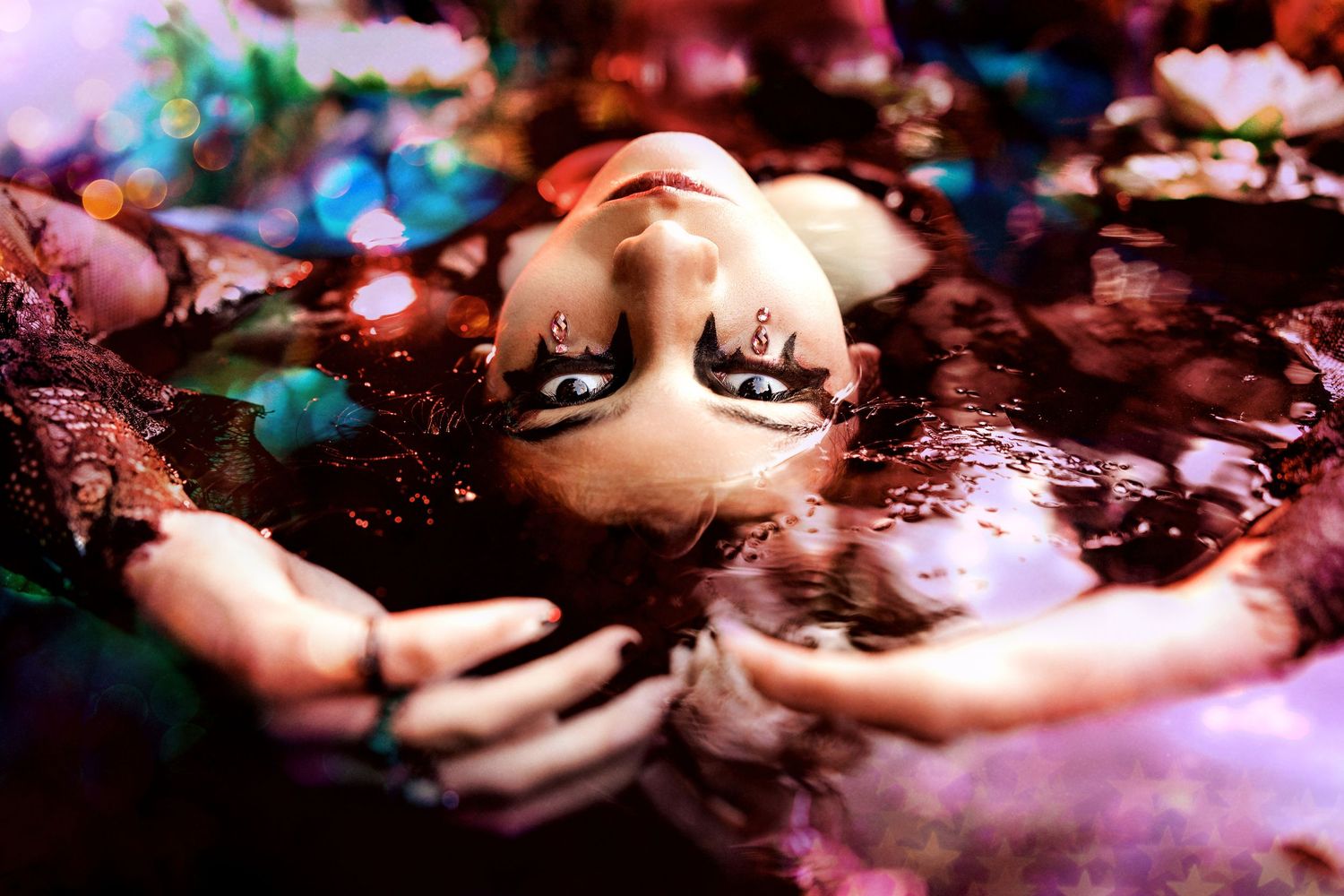 Woman with dramatic black eye makeup floats in water with colorful bokeh lights in artistic portrait.