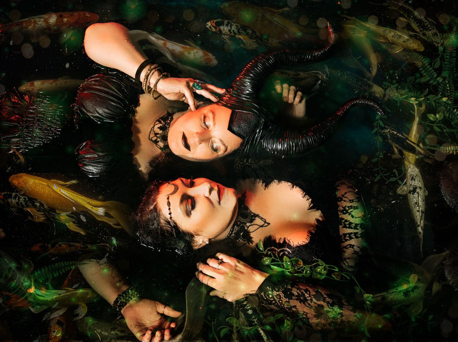 Two women in dark fantasy costumes with horns lying among colorful koi fish in mystical water.