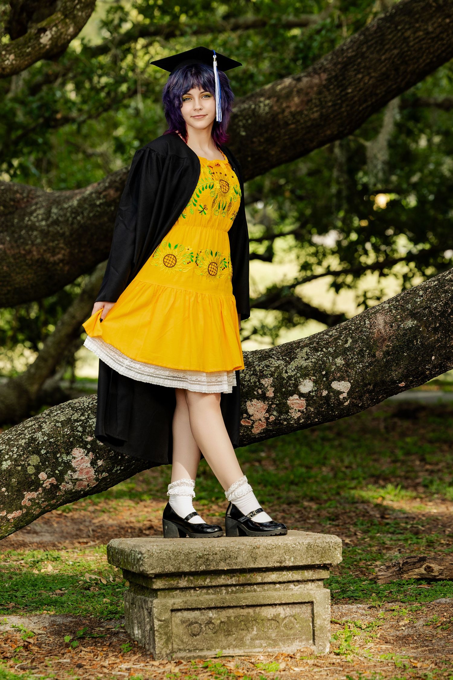 Graduate in black cap and gown with yellow sunflower dress stands on stone pedestal among trees.