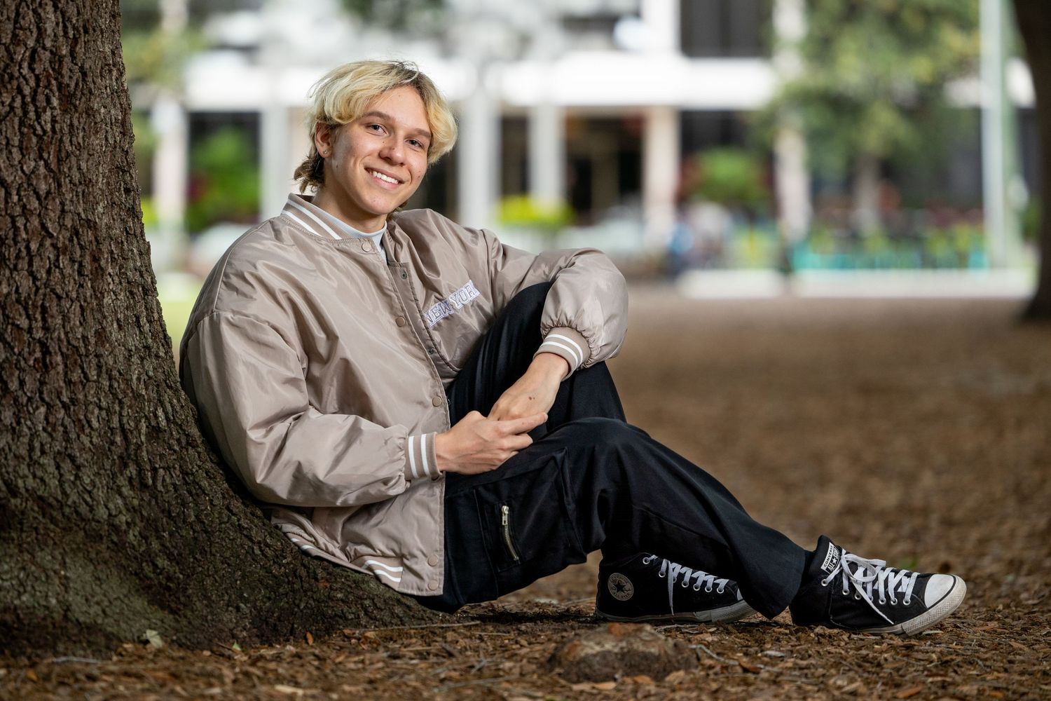 Young man with blonde hair sitting against a tree trunk outdoors, wearing a silver jacket and black jeans.