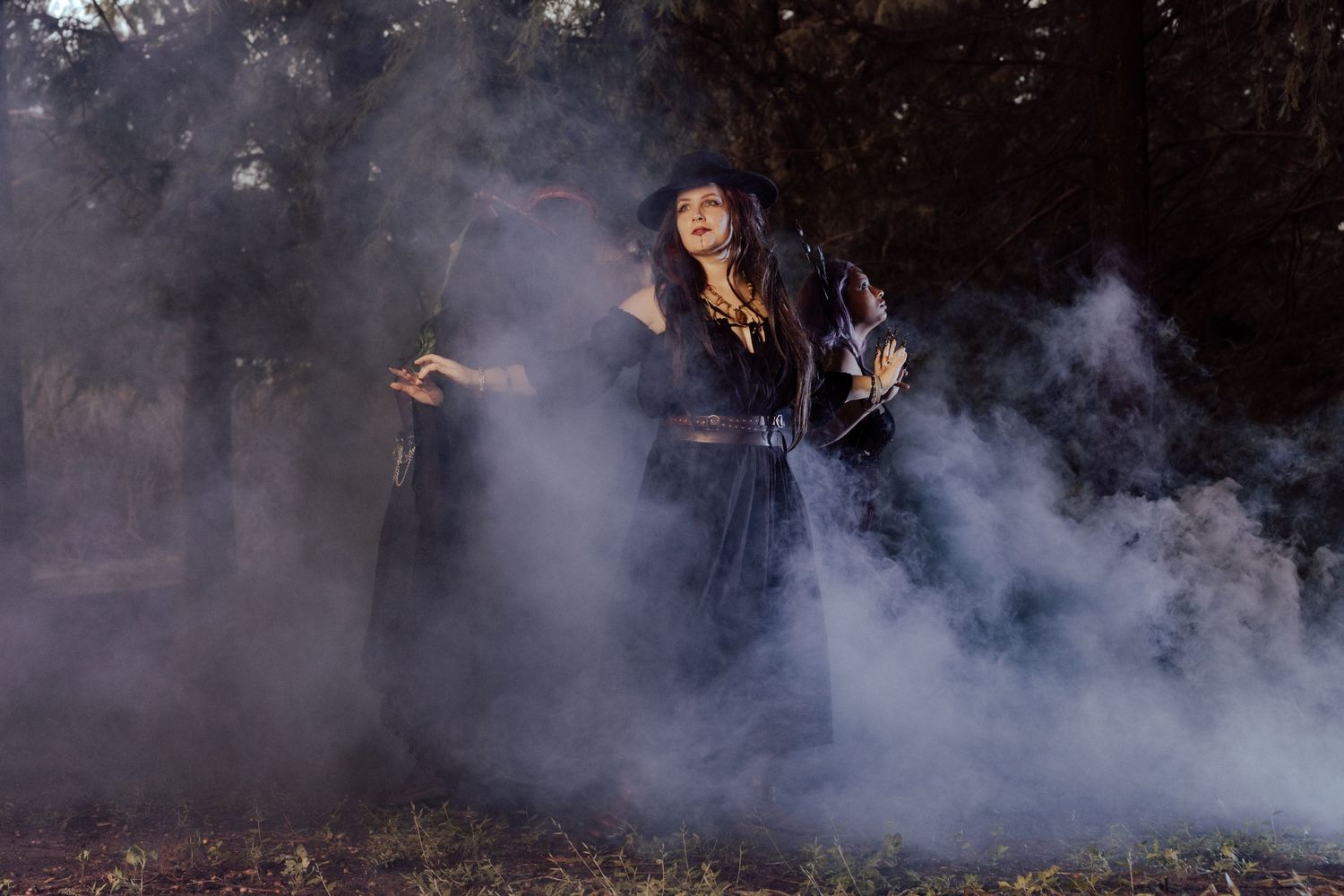 A woman in a black witch costume and wide-brim hat stands surrounded by billowing smoke in a dark forest.