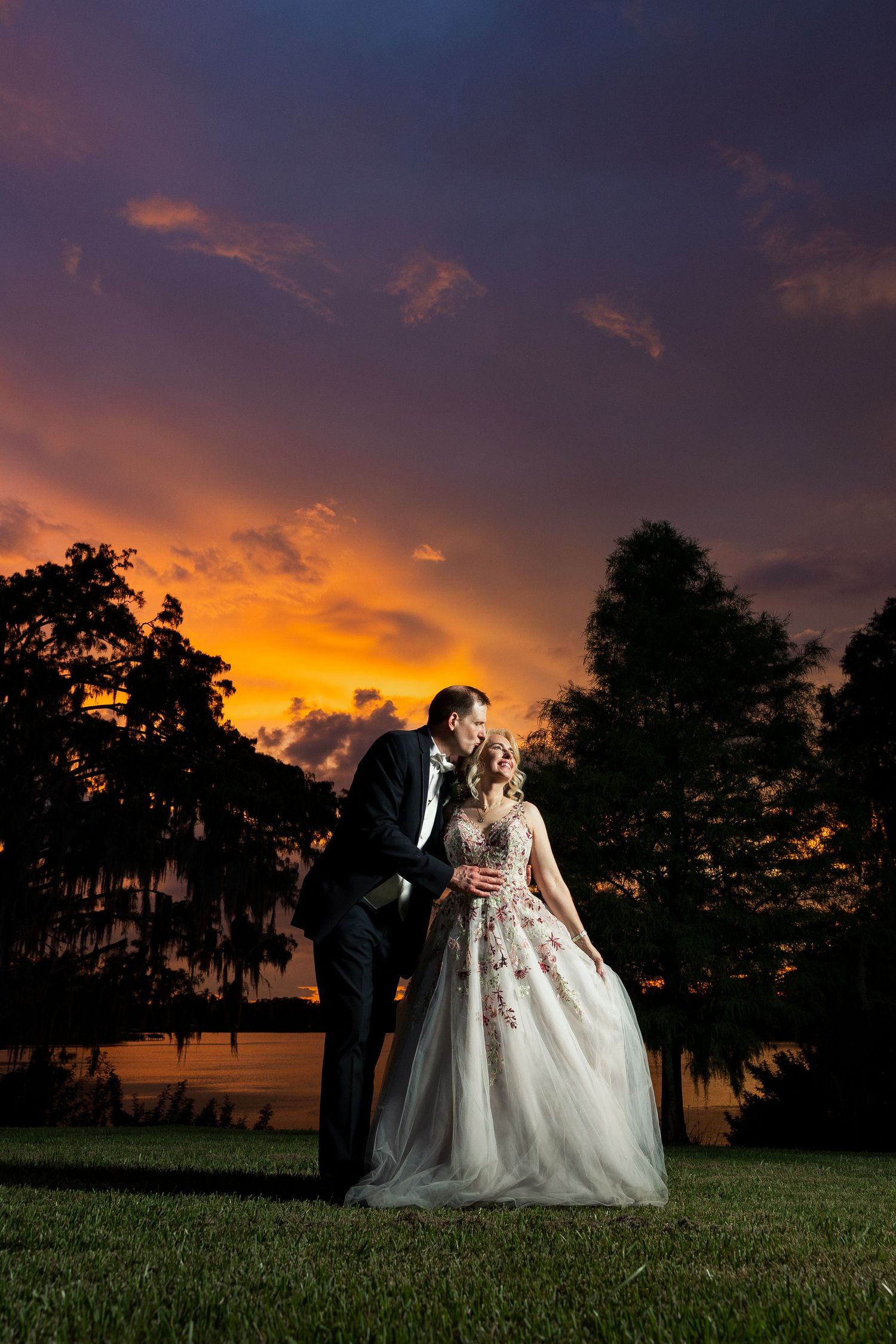 Bride and groom embrace at sunset with vibrant orange and purple sky, silhouetted trees, and a serene lake backdrop.