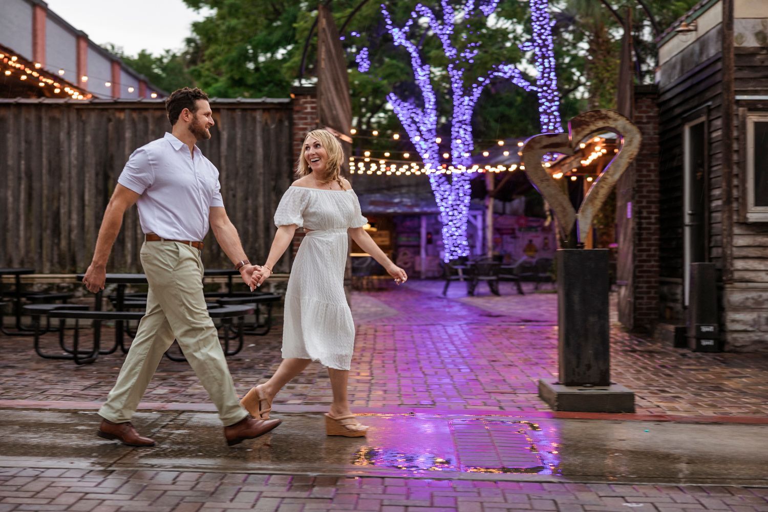 Couple walking hand in hand on wet pavement with purple lit trees in background.