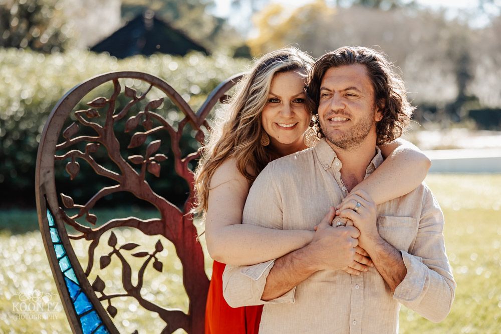 Sunny outdoor portrait of couple embracing by decorative metal bench in garden setting.