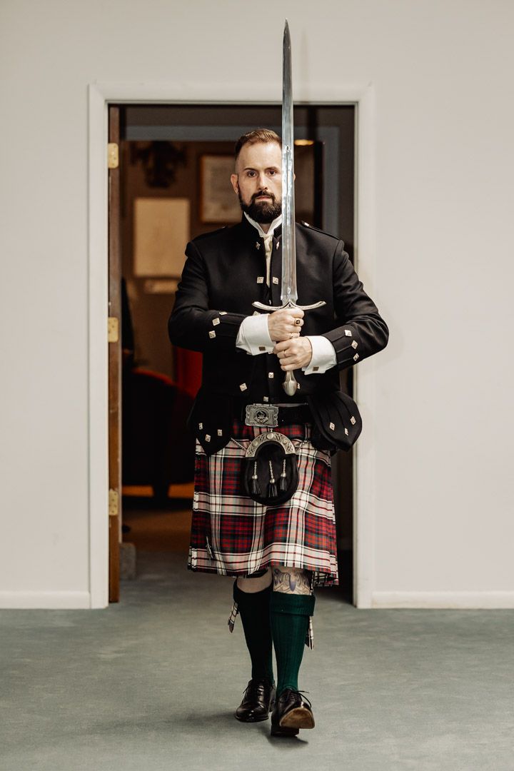 A person in traditional Scottish attire holds a ceremonial sword in a formal pose.