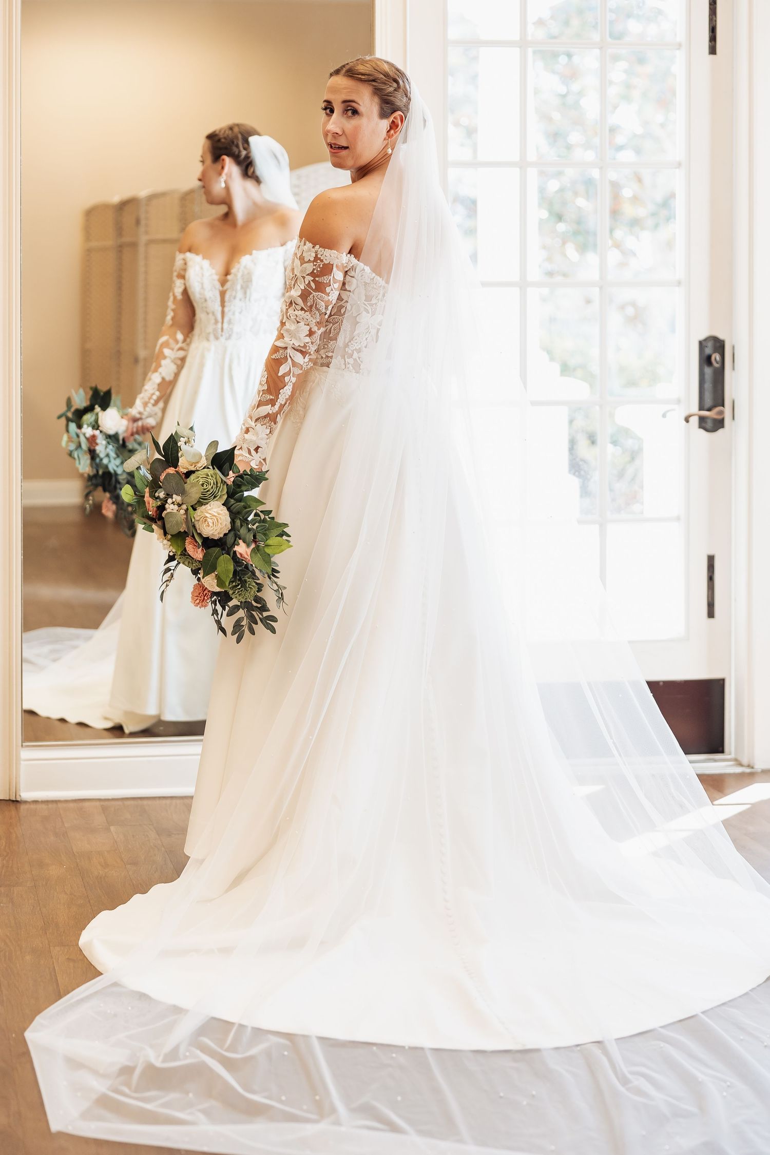 Bride in off-shoulder lace gown with long train and veil reflected in a mirror.