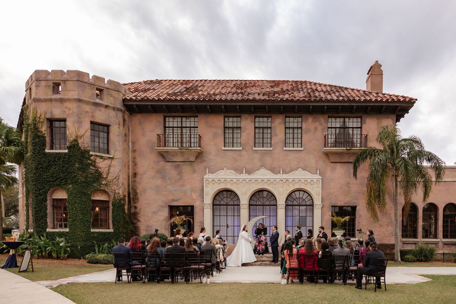 Wide shot of outdoor wedding ceremony at grand Spanish Mediterranean estate with ivy-covered walls.